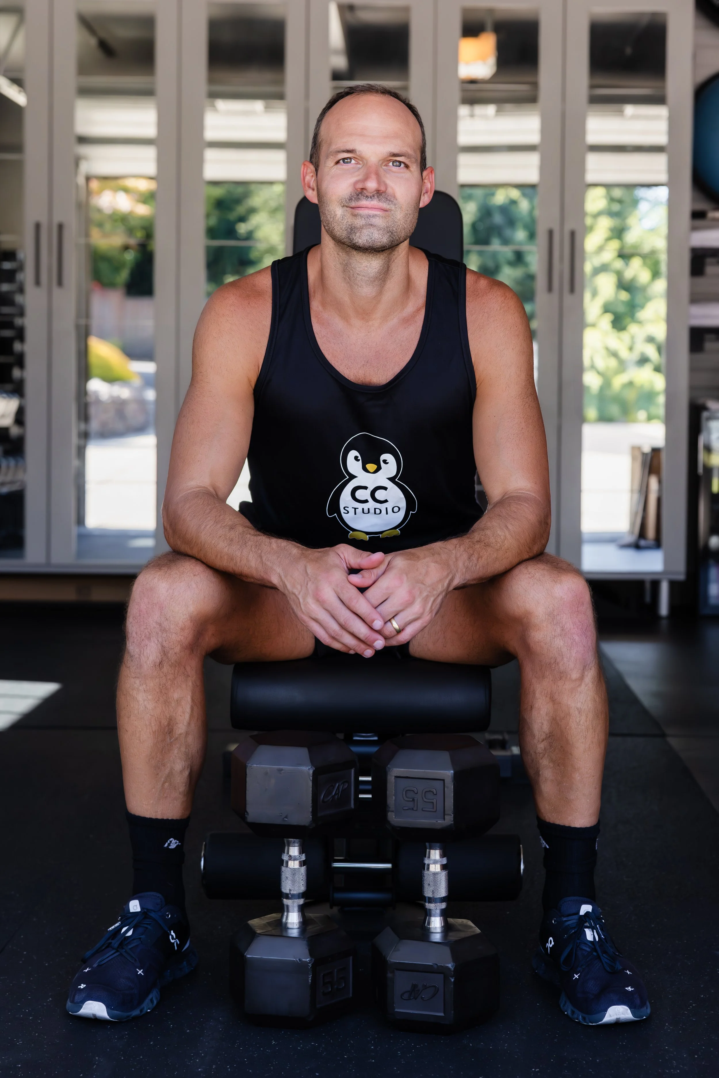 A man sits on a workout bench inside a gym, wearing a black tank top with a penguin logo and the text "CC Studio", black shorts, black sneakers, and black socks. There are dumbbells placed in front of him, and the background shows gym equipment and a window with a view of green trees.