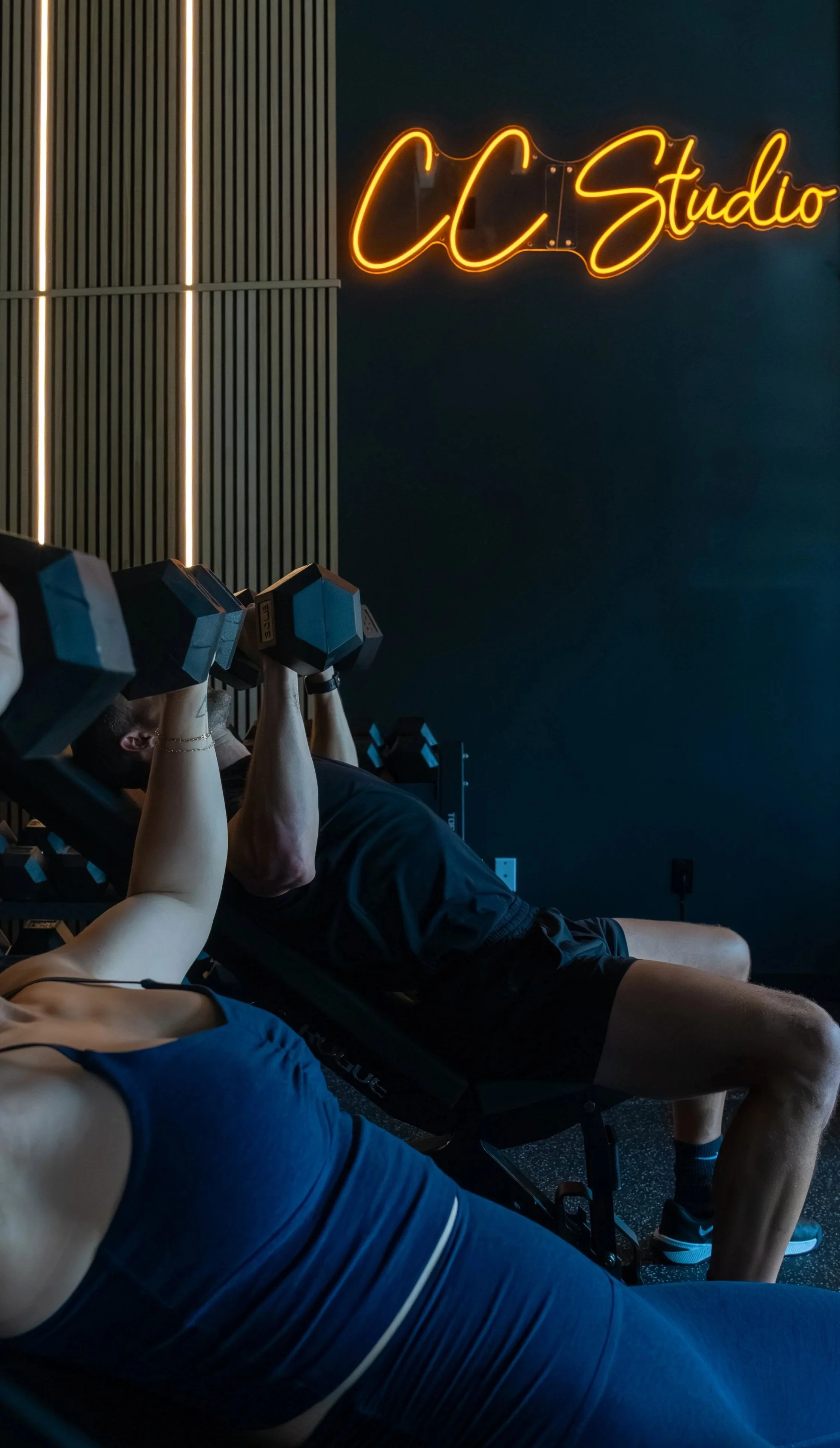People exercising with dumbbells in a gym, with a neon sign that reads 'CC Studio' on the wall.