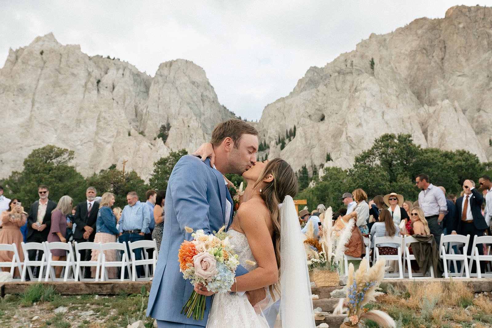 A bride and groom sharing a kiss during their outdoor wedding at the base of rocky mountains, with guests seated and standing in the background.