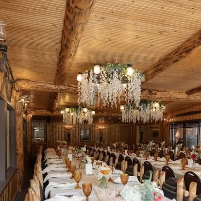 A large banquet hall decorated for an event with long tables, floral centerpieces, and elegant table settings. The ceiling features wooden beams and hanging chandeliers adorned with hanging floral arrangements. The room has wood-paneled walls and windows with curtains.