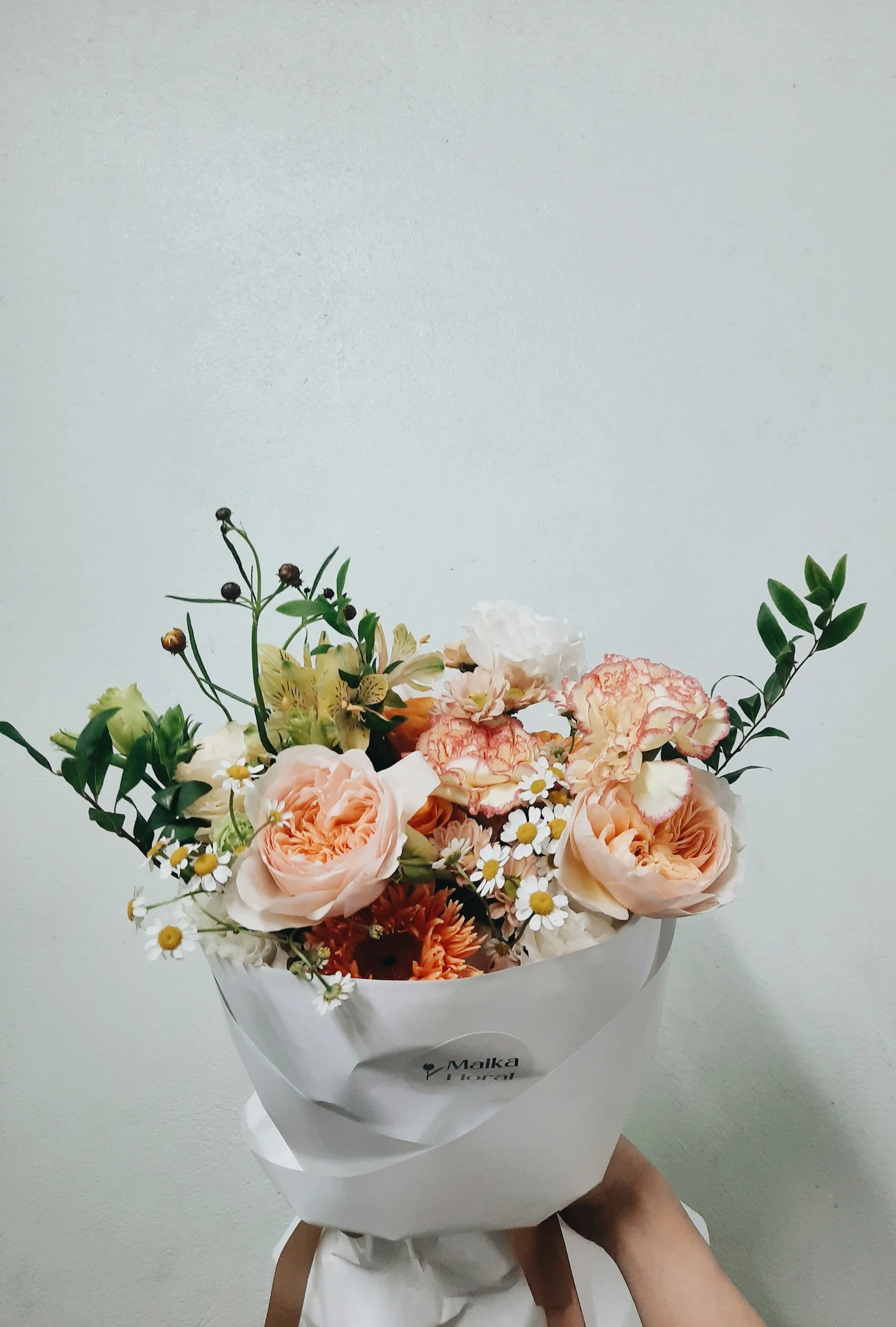 A bouquet of various flowers including roses, daisies, and carnations in shades of peach, white, and orange, wrapped in white paper with a brown ribbon, being held against a plain light-colored background.