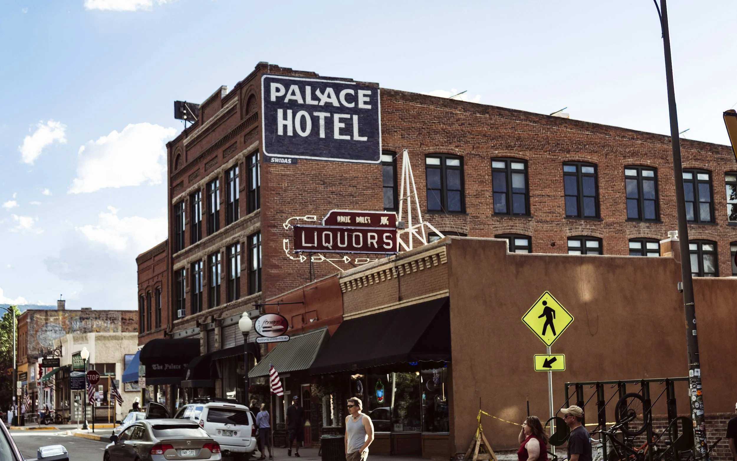 A busy street corner with a historic brick building featuring signs for Palace Hotel and Liquors, pedestrians walking, bicycles, and a yellow pedestrian crossing sign.