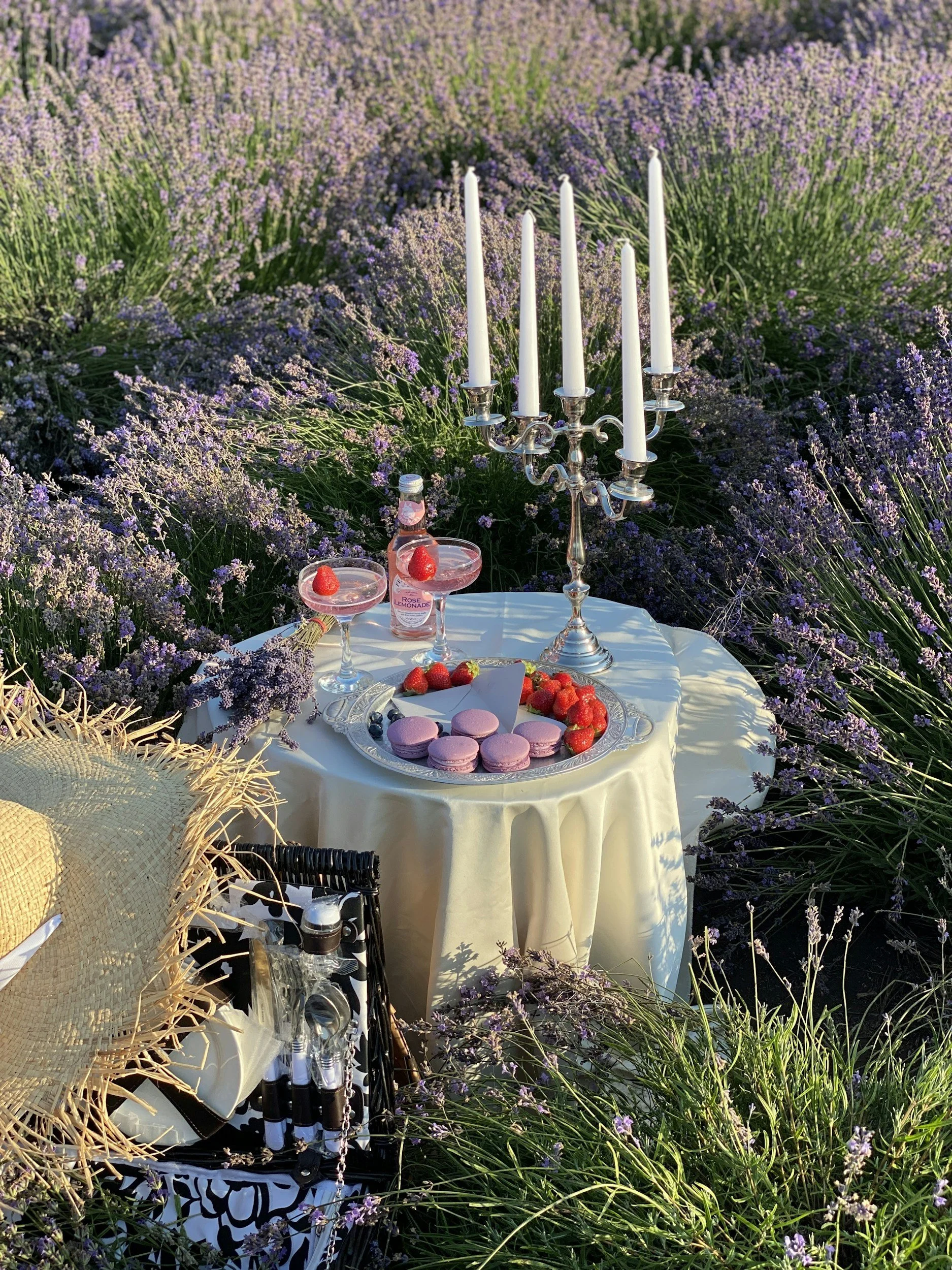 A picnic setup with a white tablecloth-covered table surrounded by lavender flowers, featuring a silver candelabrum with seven unlit candles, two pink cocktails with strawberries, a bottle of rose lemonade, pink macarons, strawberries, blueberries, a straw hat, and picnic utensils in a basket.