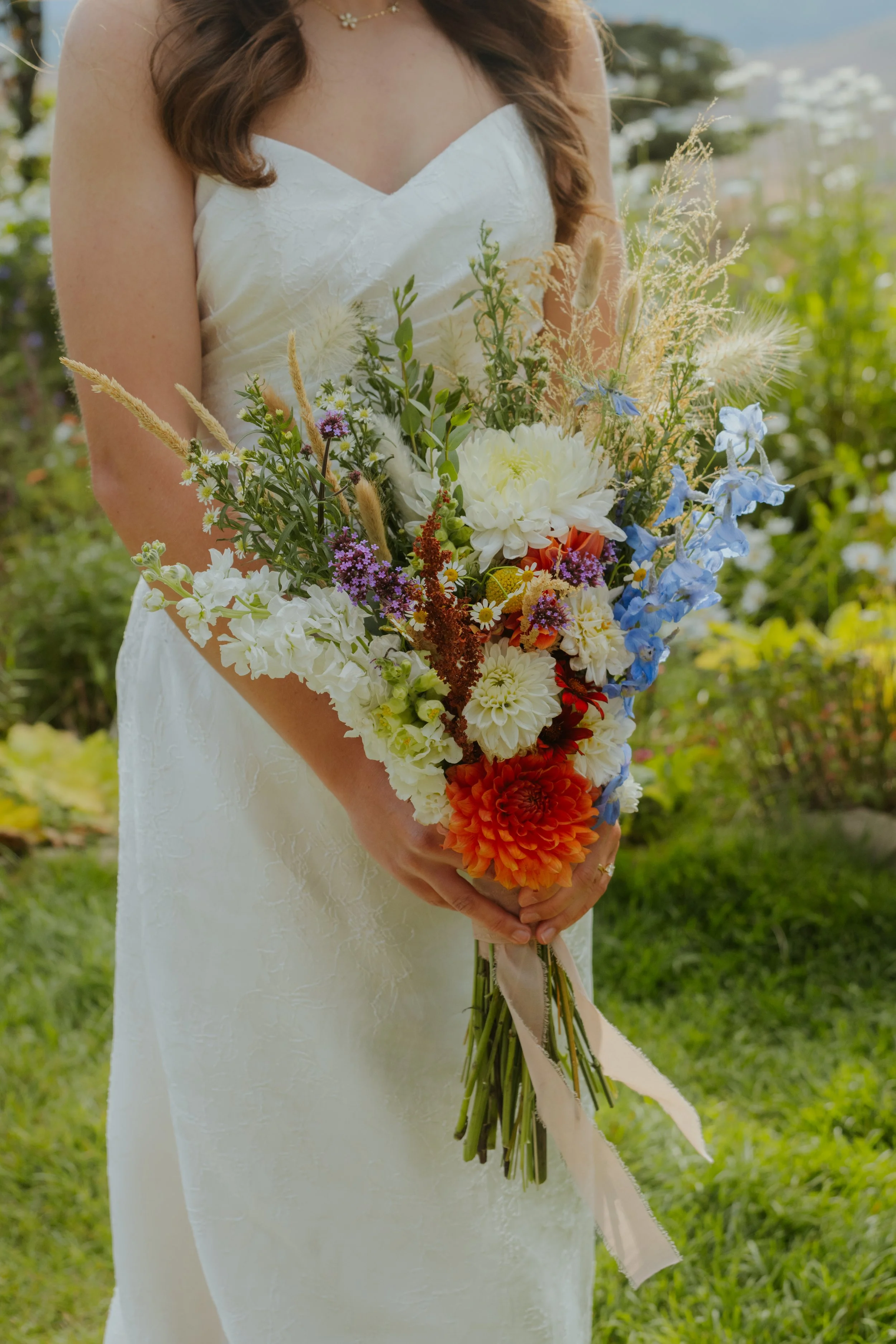 Close-up of a bride holding a colorful bouquet of flowers outdoors, with a garden and buildings in the background.
