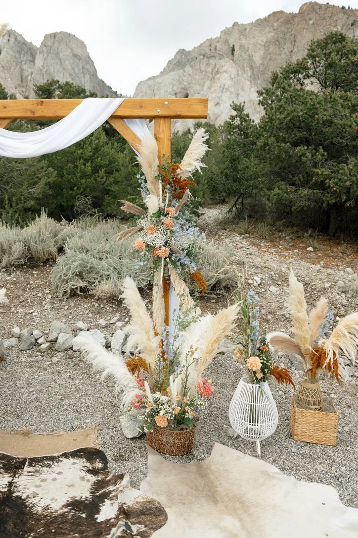 A wooden wedding arbor decorated with white fabric and a floral arrangement of pampas grass, roses, and greenery, set outdoors against a mountain landscape.