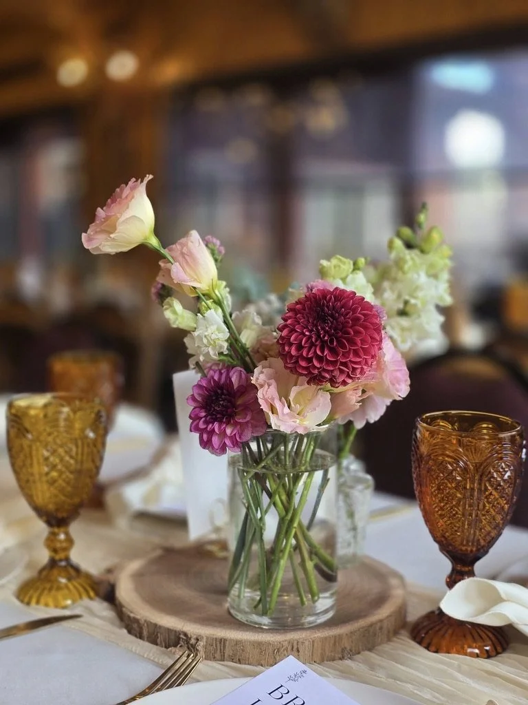 A floral centerpiece with pink, purple, and white flowers in a glass vase on a wooden slab atop a table with amber-colored glassware and a white tablecloth.