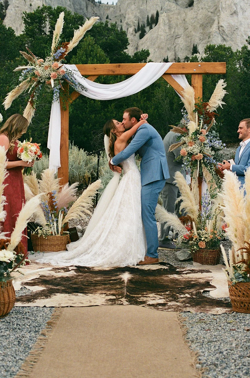 A bride and groom sharing a kiss during their outdoor wedding ceremony at Deer Valley Ranch under a wooden arch decorated with flowers and white fabric, with guests and natural landscape in the background.