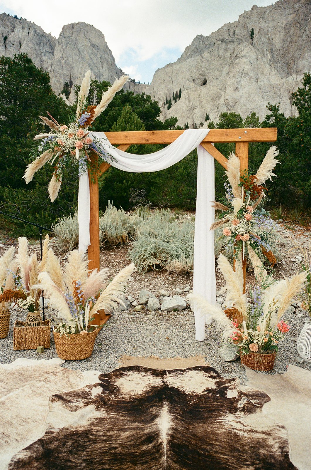 Boho wedding arch installation at Deer Valley Ranch in Salida, Colorado — fresh peach and dusty blue florals, pampas grass, boho grasses, white linen drape on a wooden ceremony arch with a granite mountain backdrop. Full ground installation included.