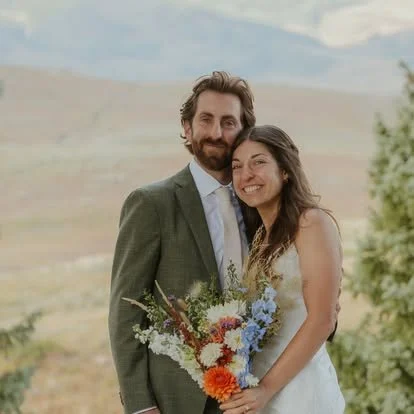 A man and a woman smiling, holding a bouquet of flowers, outdoors with a scenic landscape background.