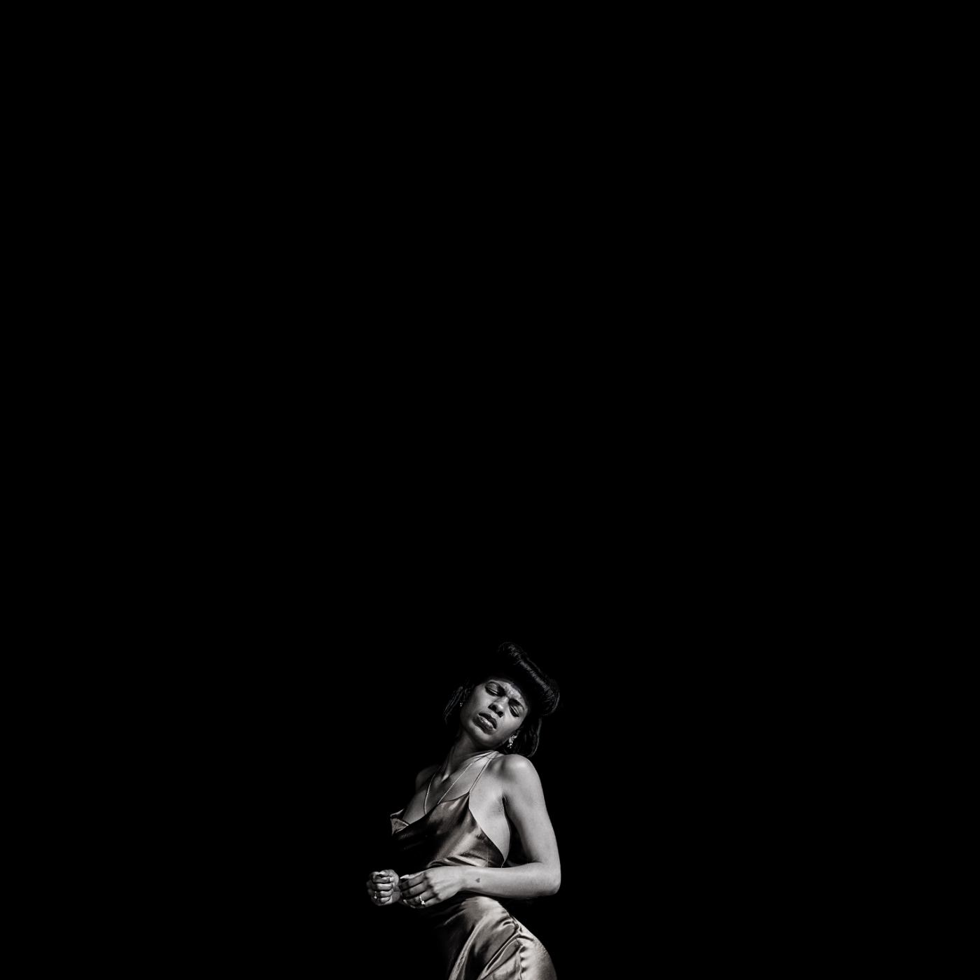 Black and white photo of a woman in a silky dress, standing against a dark background, with her eyes closed and a sensual expression.