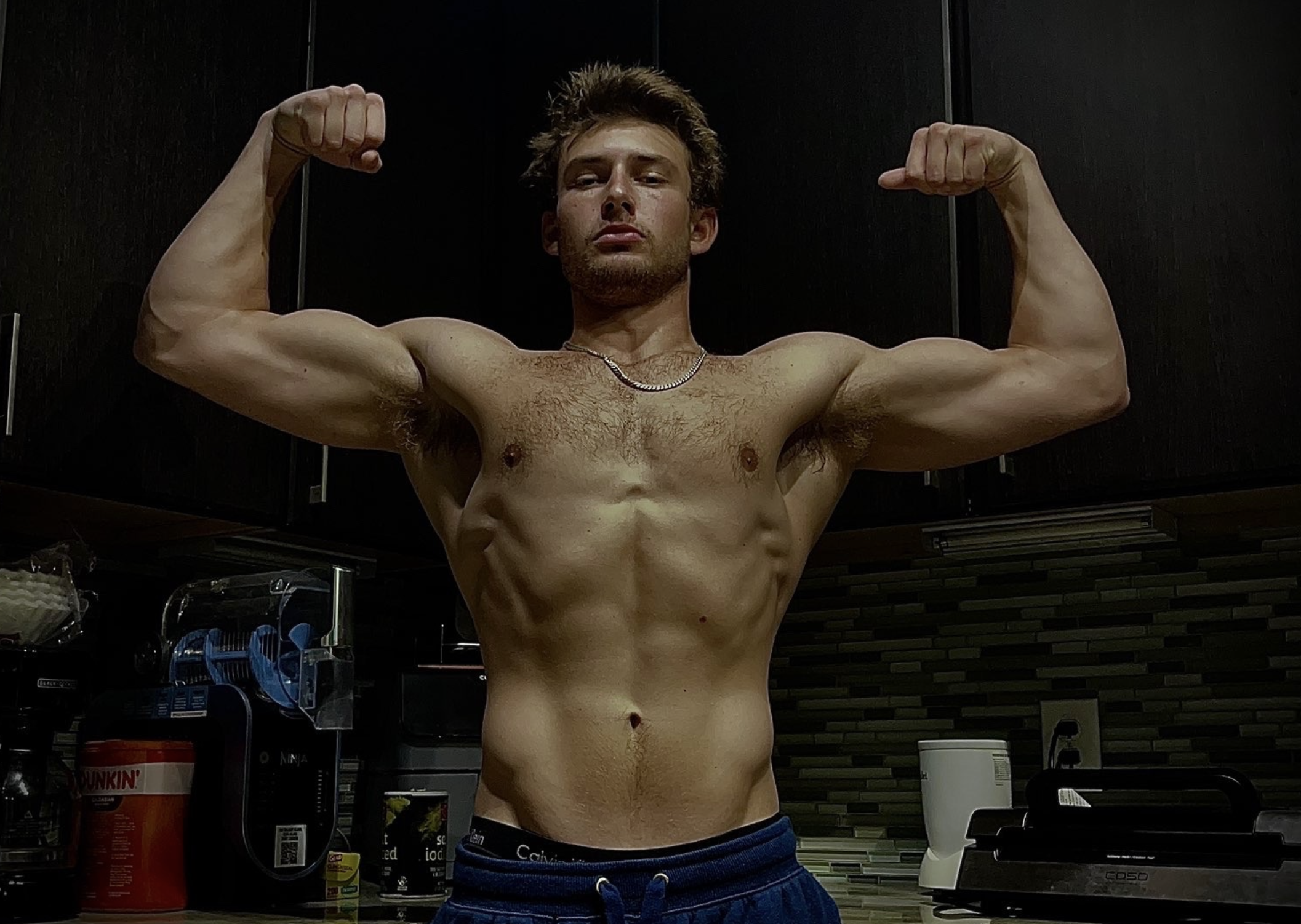 Young man with muscular physique flexing his biceps in a kitchen, shirtless, wearing blue shorts, with dark cabinets and kitchen appliances in the background.