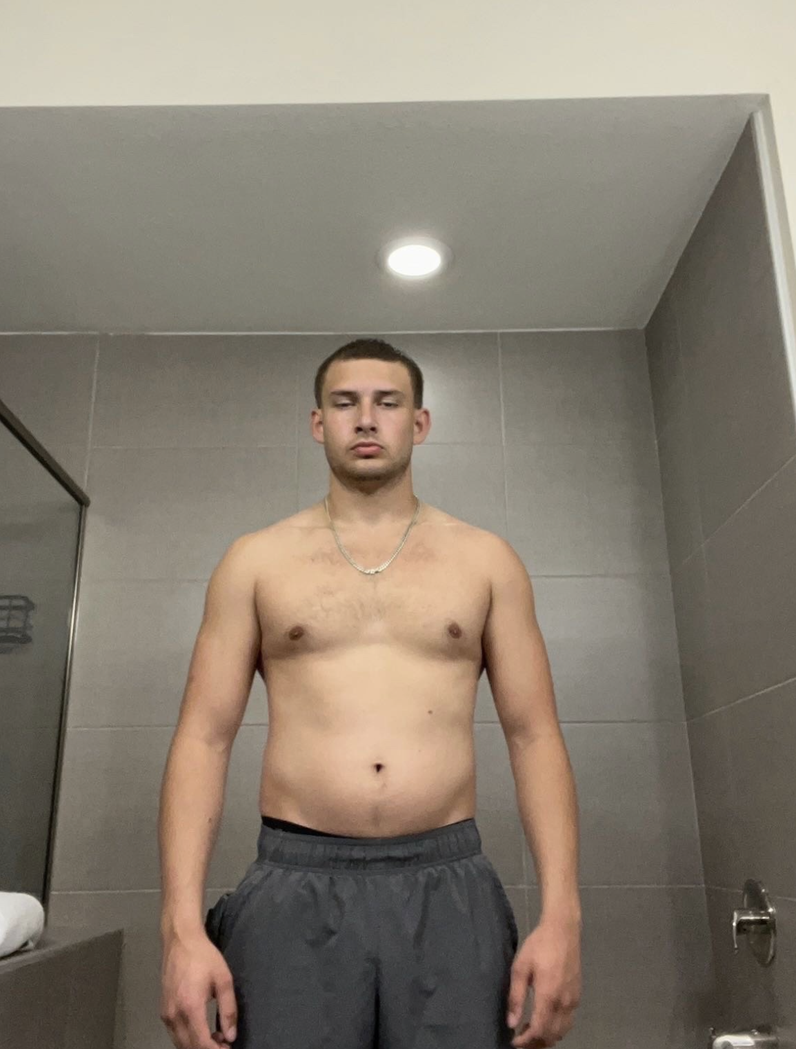 A shirtless young man standing in a bathroom, looking at the camera with a neutral expression, wearing gray shorts and a silver chain necklace.