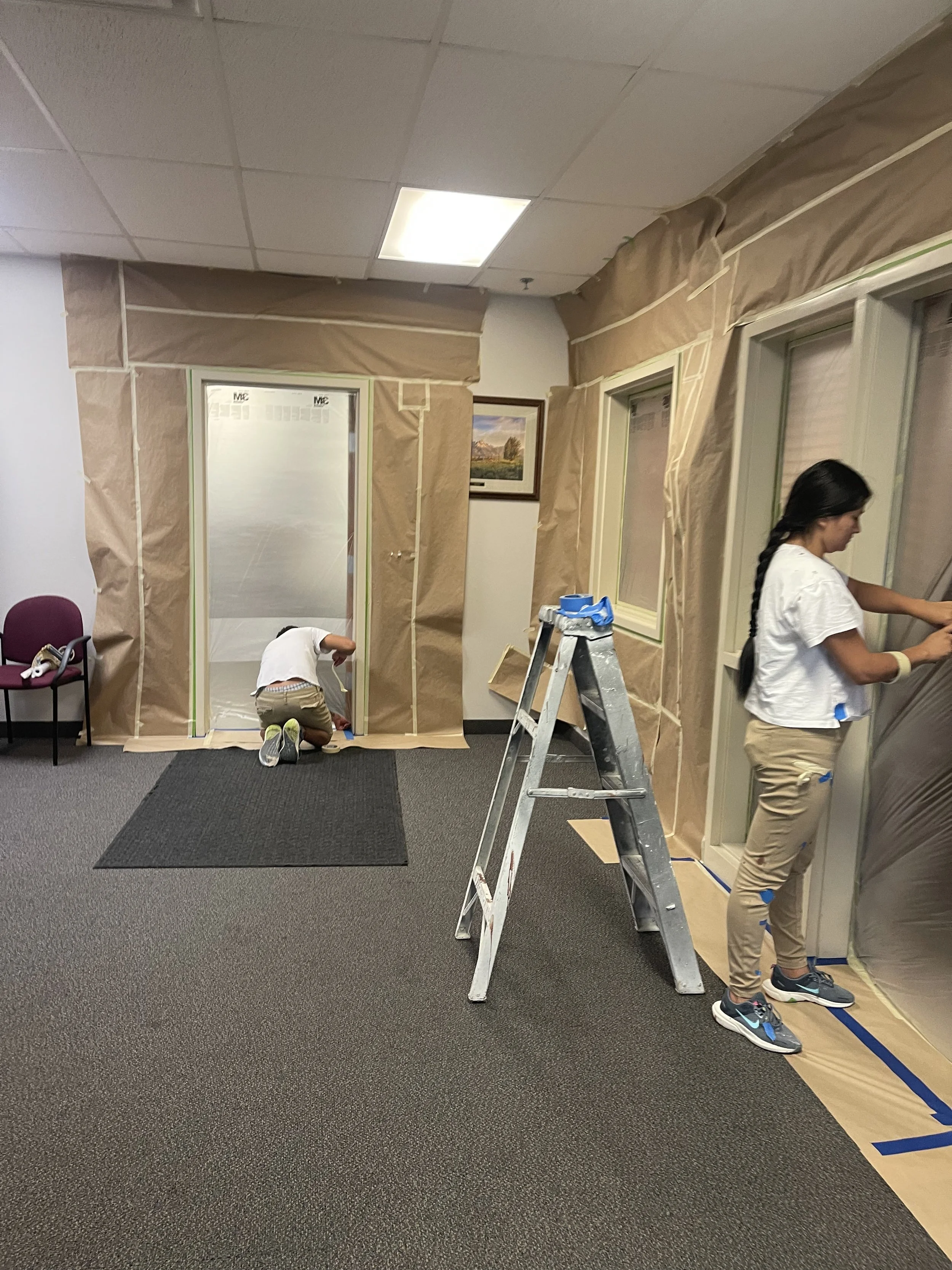 Two women are painting and preparing a room, with brown paper covering the walls and window frames, one woman kneeling near the door and the other standing by the window, working. A ladder is positioned in the middle of the room.