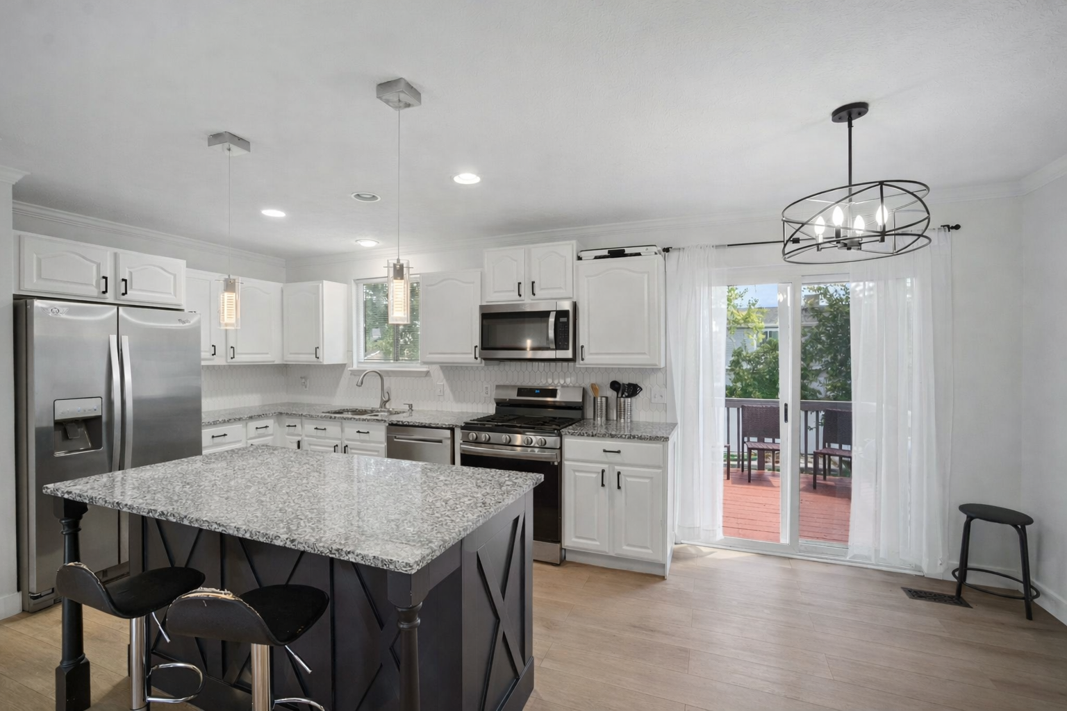 Modern kitchen with white cabinets, stainless steel refrigerator and appliances, granite countertops, island with black base, and sliding glass door leading to an outdoor patio with seating.