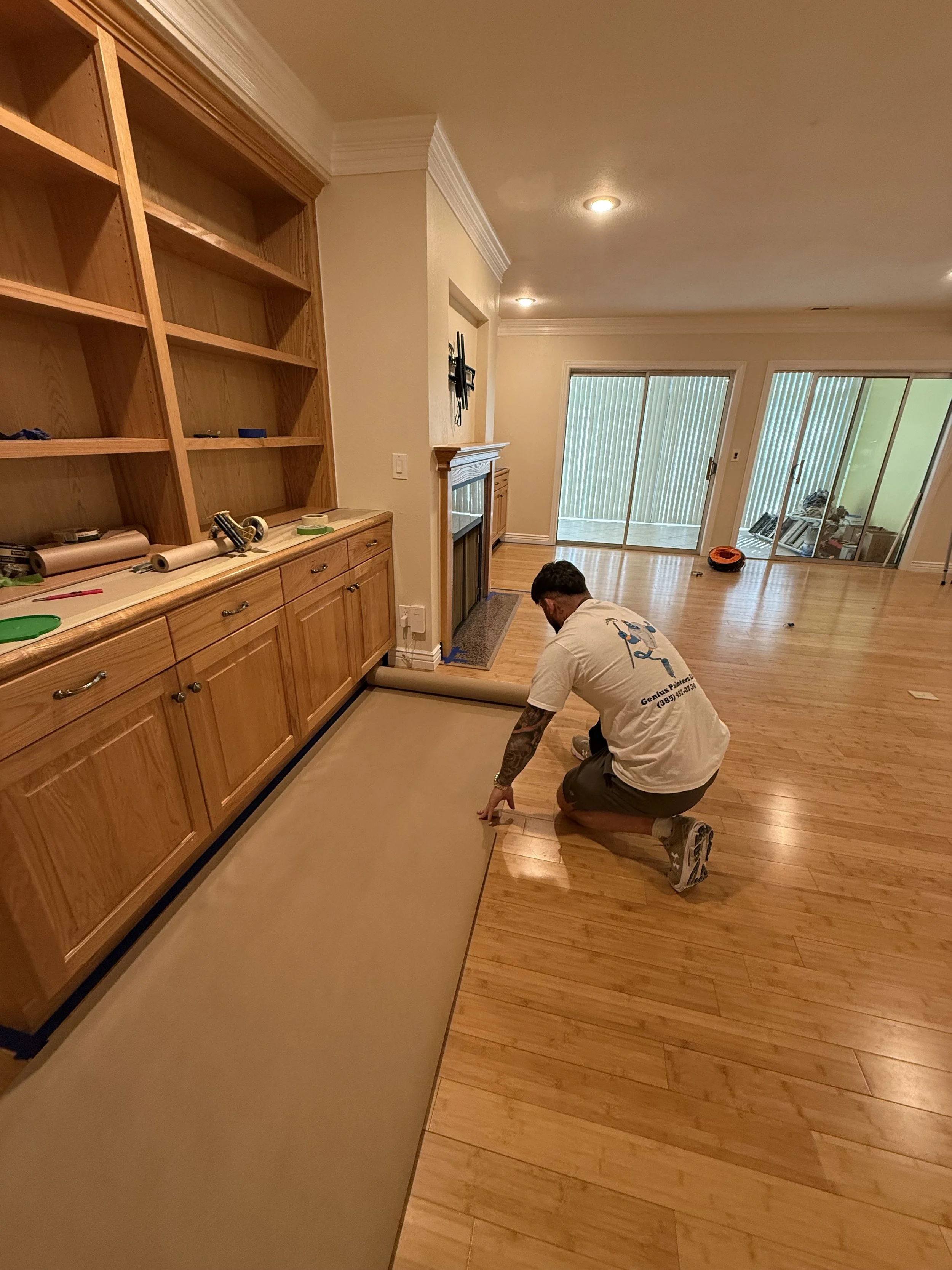 A man kneeling on a hardwood floor, unrolling a beige carpet in a living room area with large sliding glass doors leading outside.