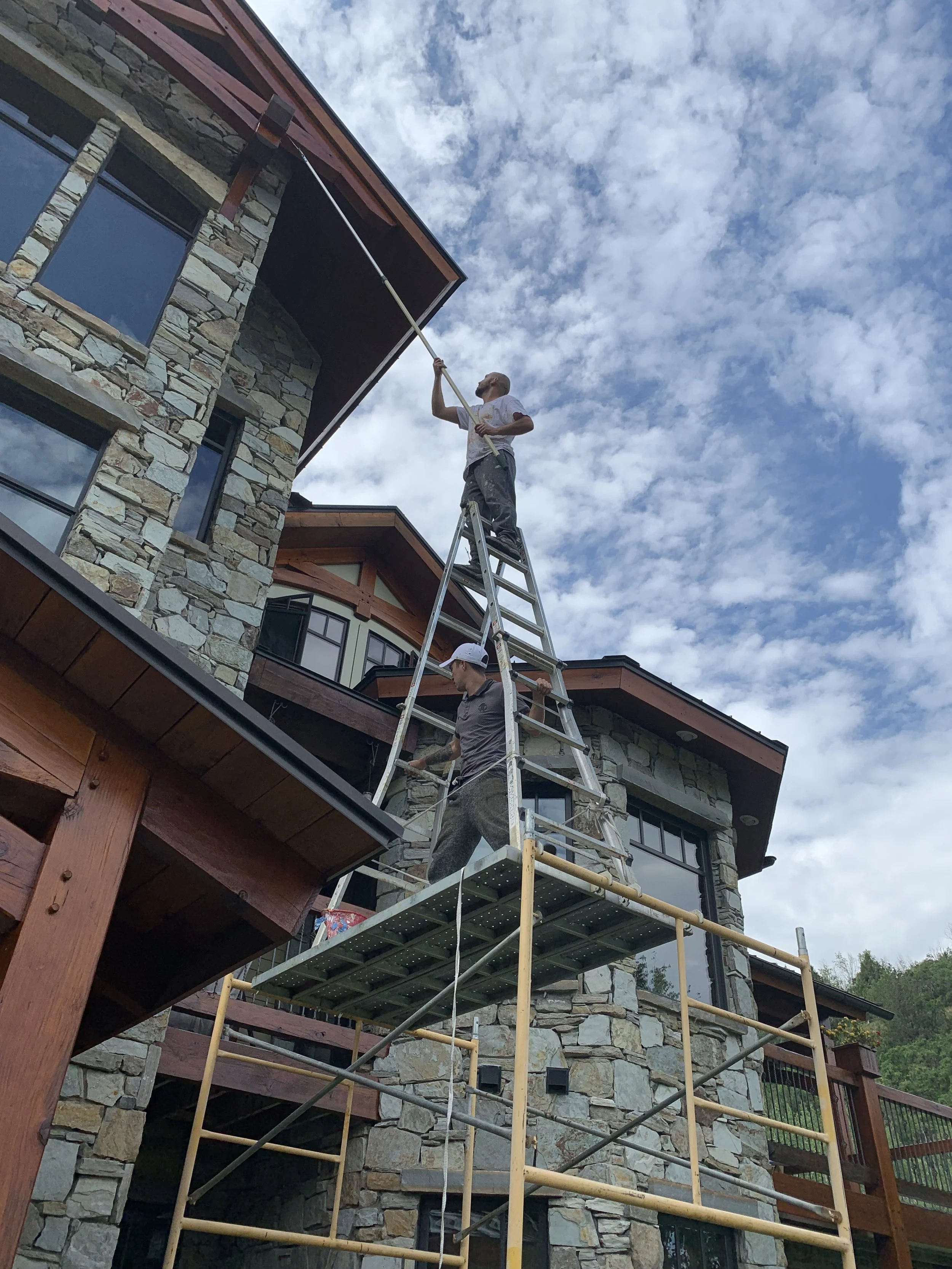 Two workers on scaffolding cleaning the exterior of a stone house with a long-handled brush, under a partly cloudy sky.