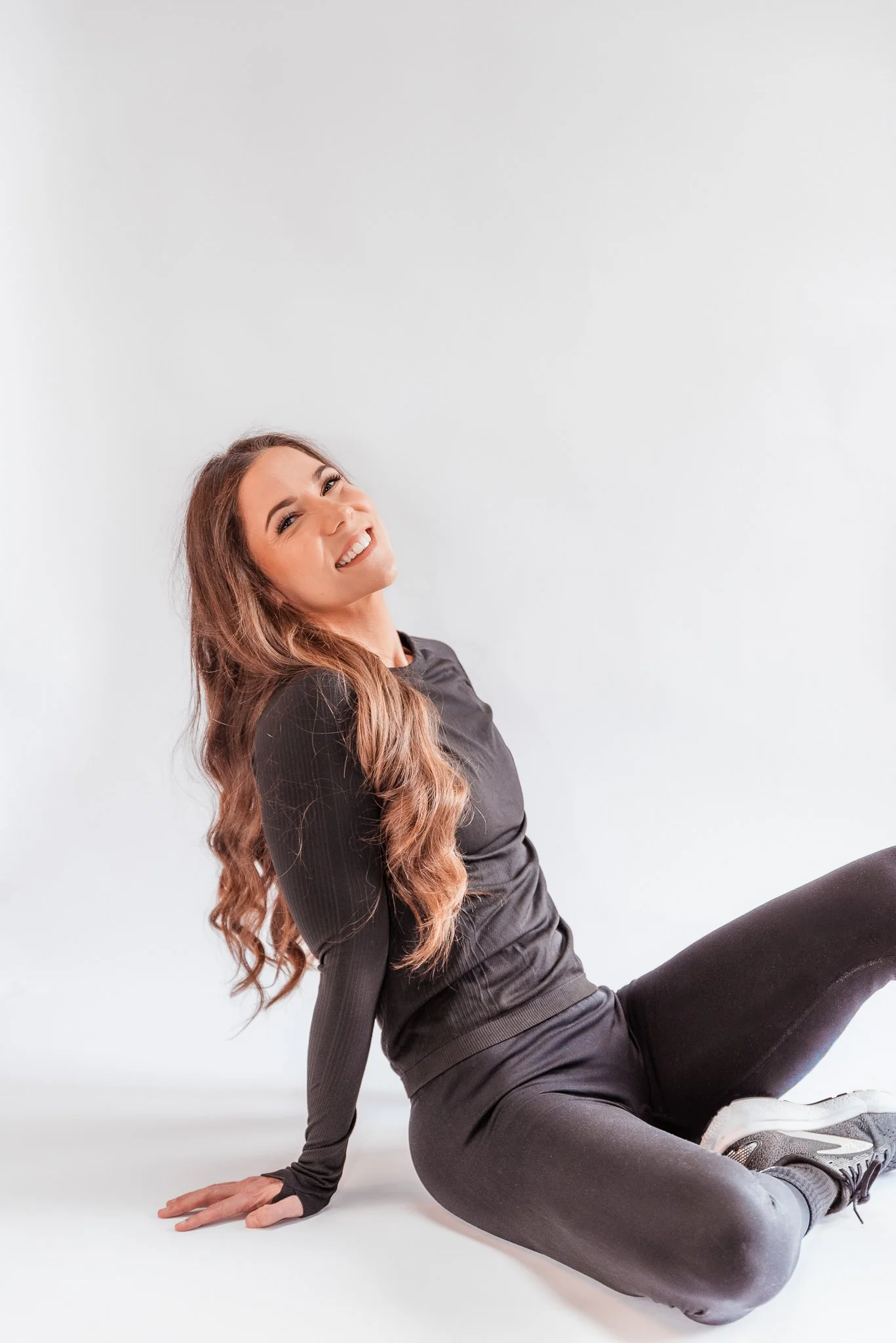 A woman with long wavy brown hair, wearing black athletic clothing and sneakers, sitting on the floor, smiling, against a plain white background.