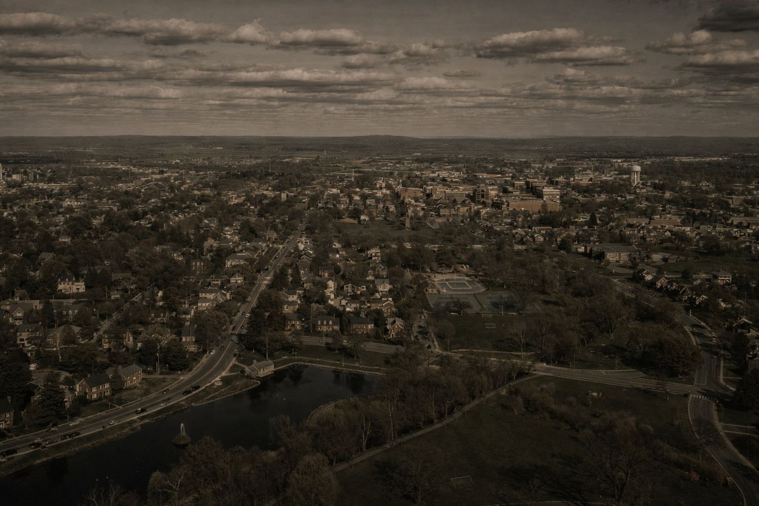 Aerial view of a city with streets, houses, a pond with a fountain, parks, and a distant skyline with a water tower.