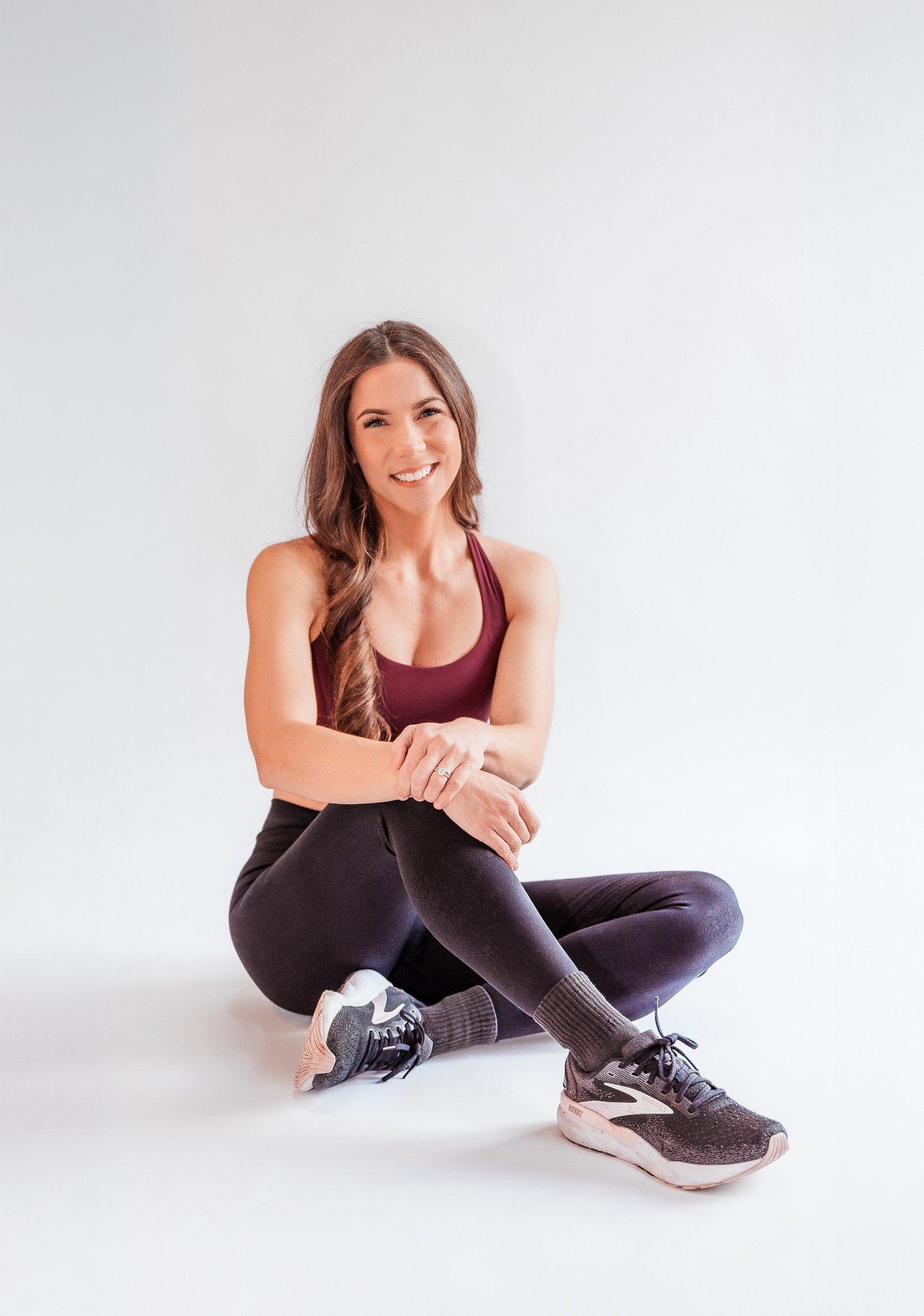 A woman sitting on the floor in activewear, smiling and looking at the camera, against a plain white background.