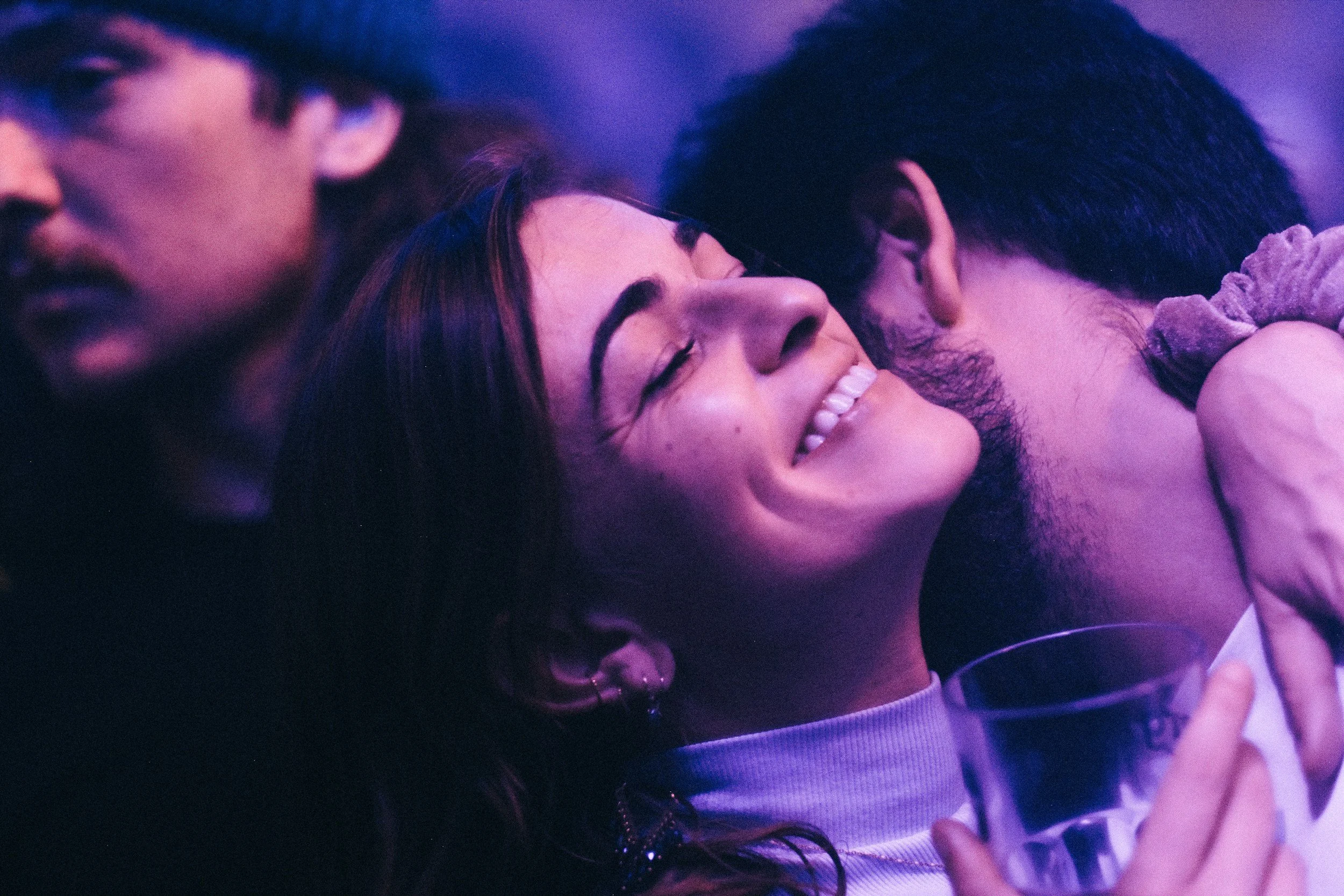 Decorative floral arch with white and pink flowers in planters, set outdoors on green grass in front of a weathered wooden fence, with white chairs flanking the aisle.
