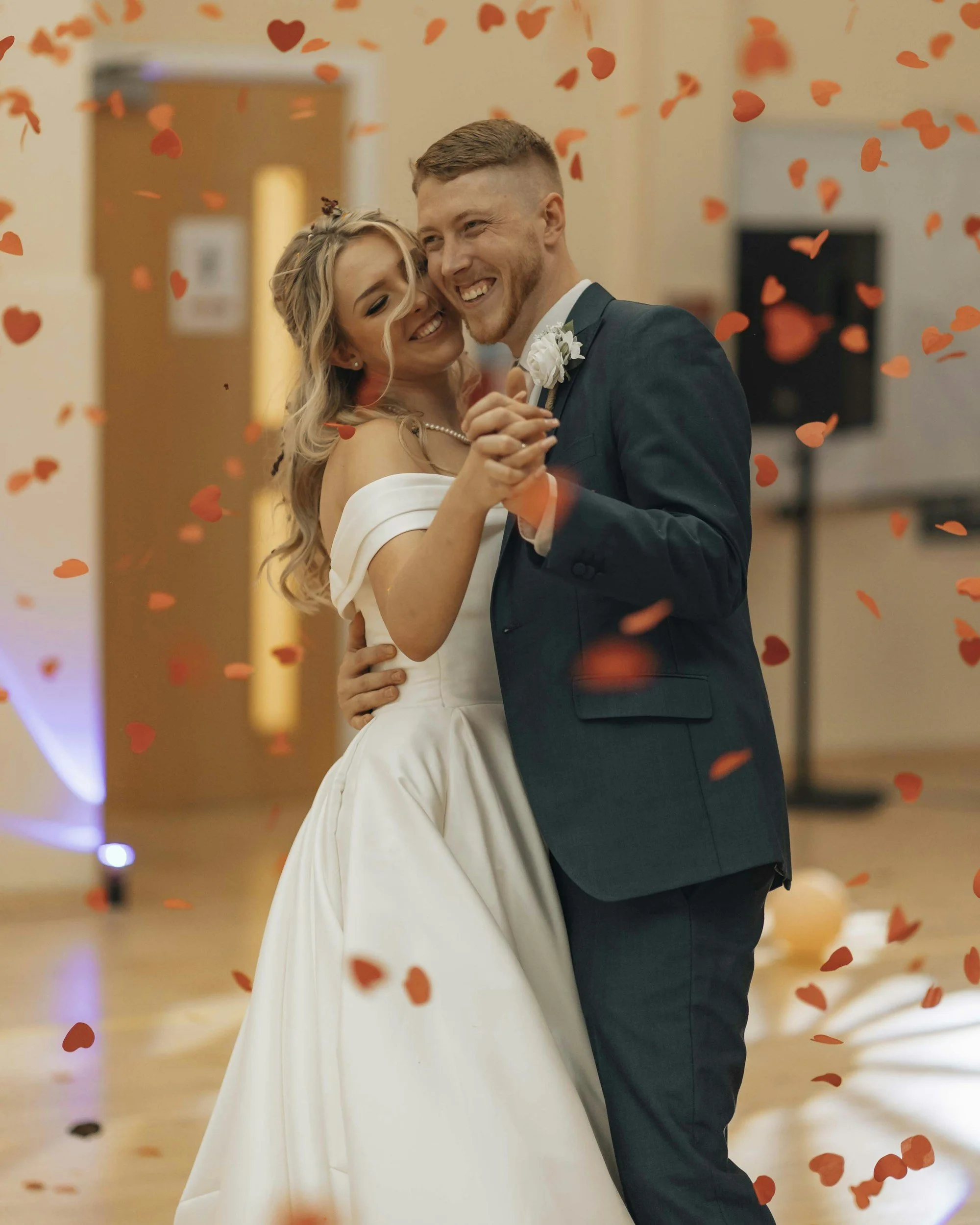 Wedding cake with a bride and groom topper flanked by floral arrangements, two glasses of champagne, plates, and cutlery on a table, against a patterned wallpaper background.
