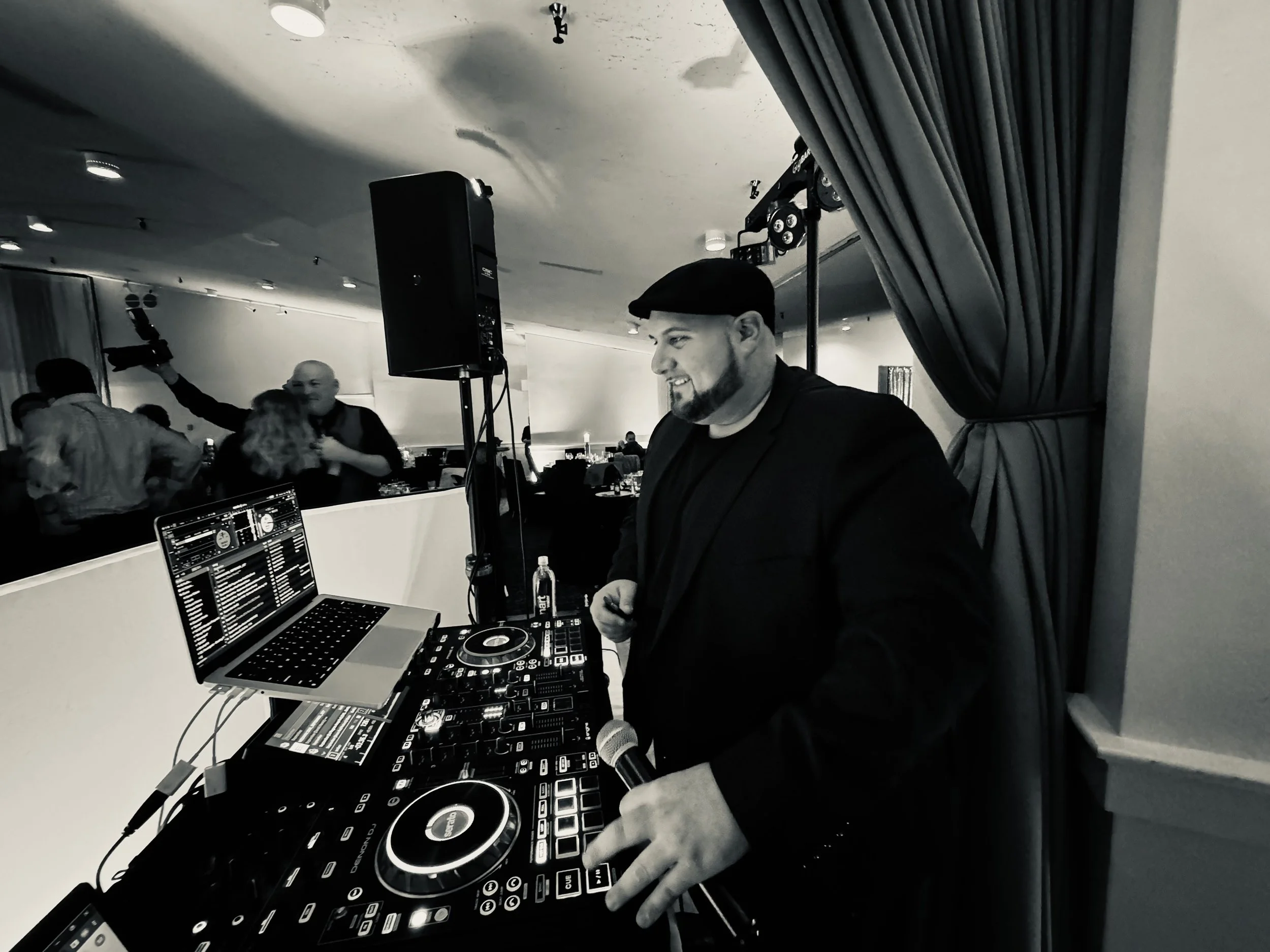 A DJ wearing a black jacket and beret, smiling and holding a microphone, stands behind a DJ booth with turntables, a laptop, and audio equipment in a dimly lit club or event space with people dancing in the background.
