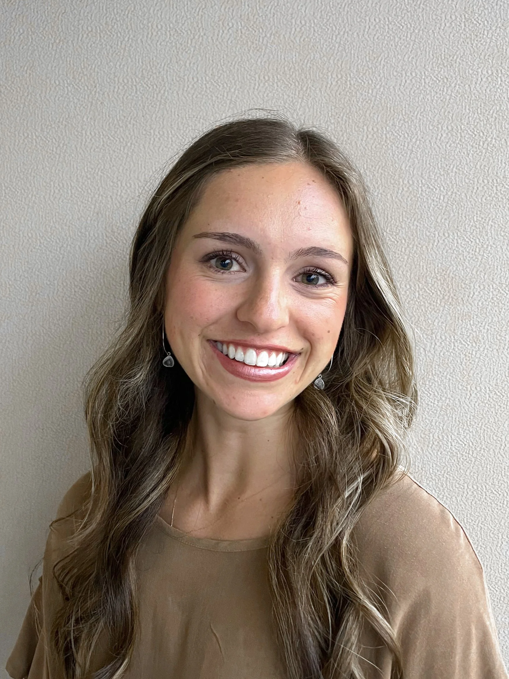 A woman with long wavy brown hair, blue eyes, and a bright smile, wearing earrings and a beige top, standing against a plain textured wall.