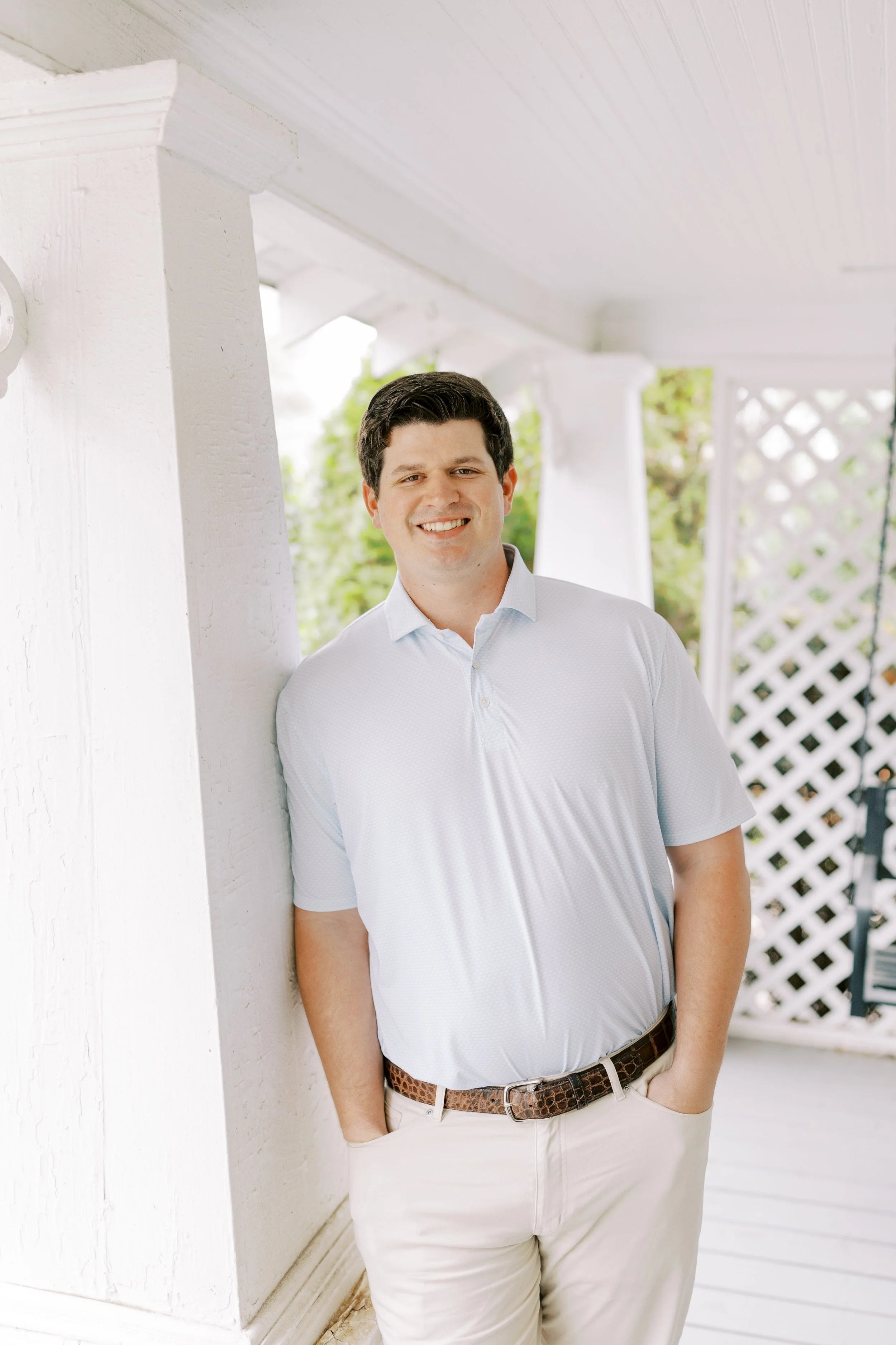 A smiling man in a white polo shirt and beige pants, with his hands in his pockets, leaning against a white wooden post on a porch with lattice and trees in the background.