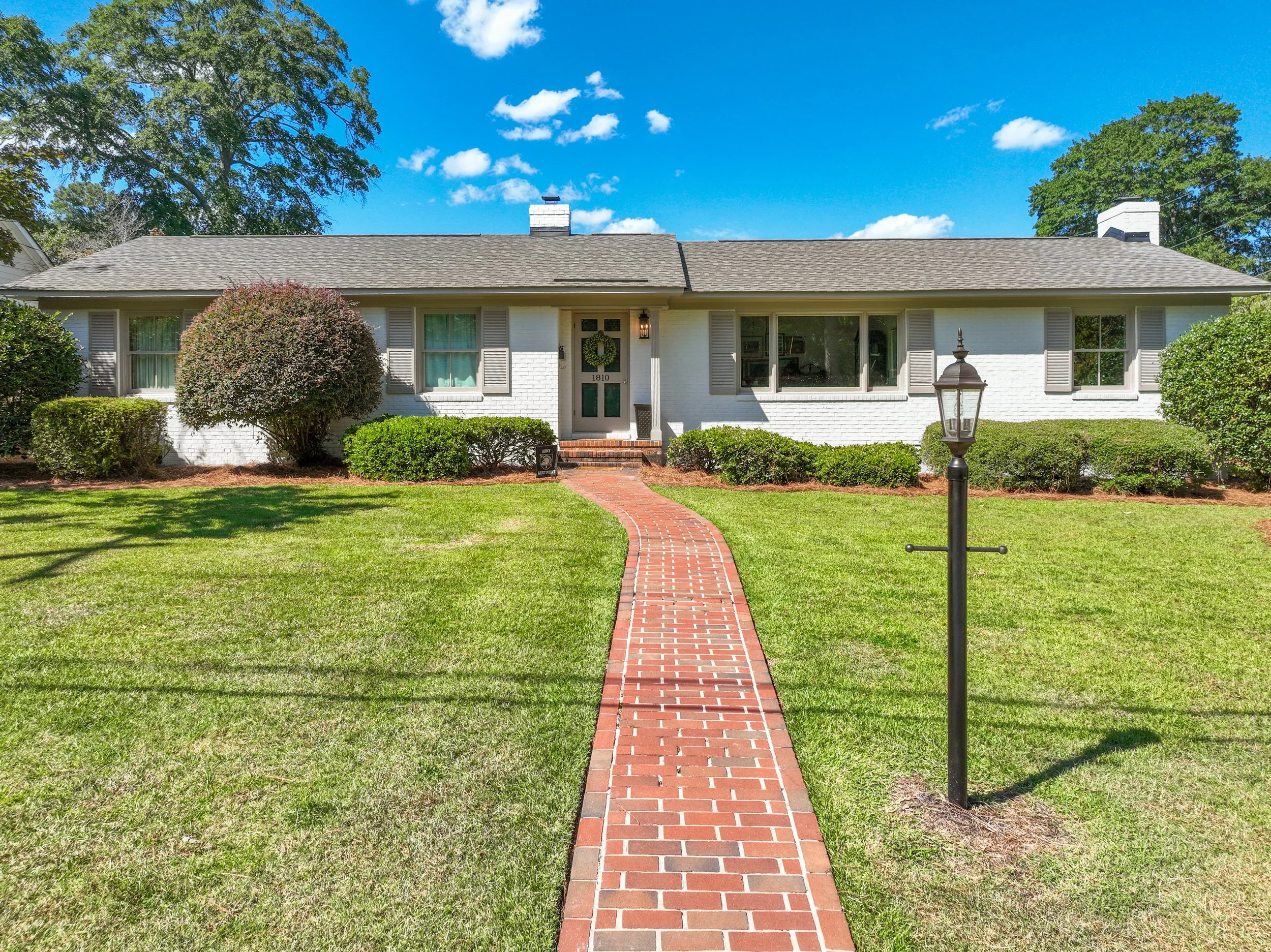 White brick house with a brick pathway leading to the front door, surrounded by well-manicured lawn and shrubs, under a bright blue sky with scattered clouds.