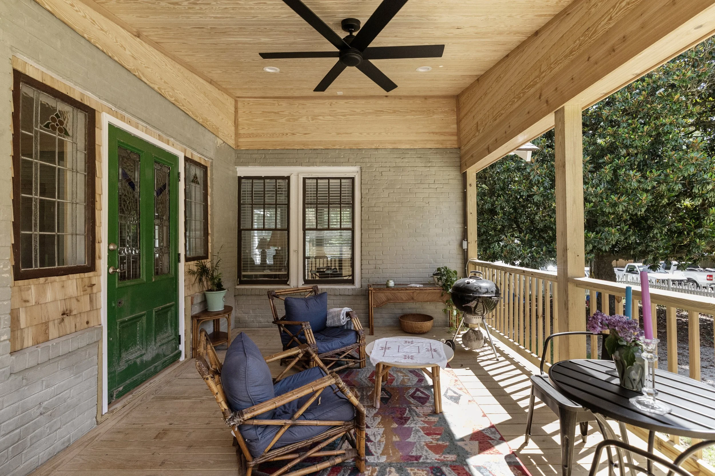 Covered porch with wooden flooring and railing, featuring outdoor furniture, a hammock chair, a small table, plants, a barbecue grill, and a table with candles. There is a ceiling fan, two windows, and a green door.