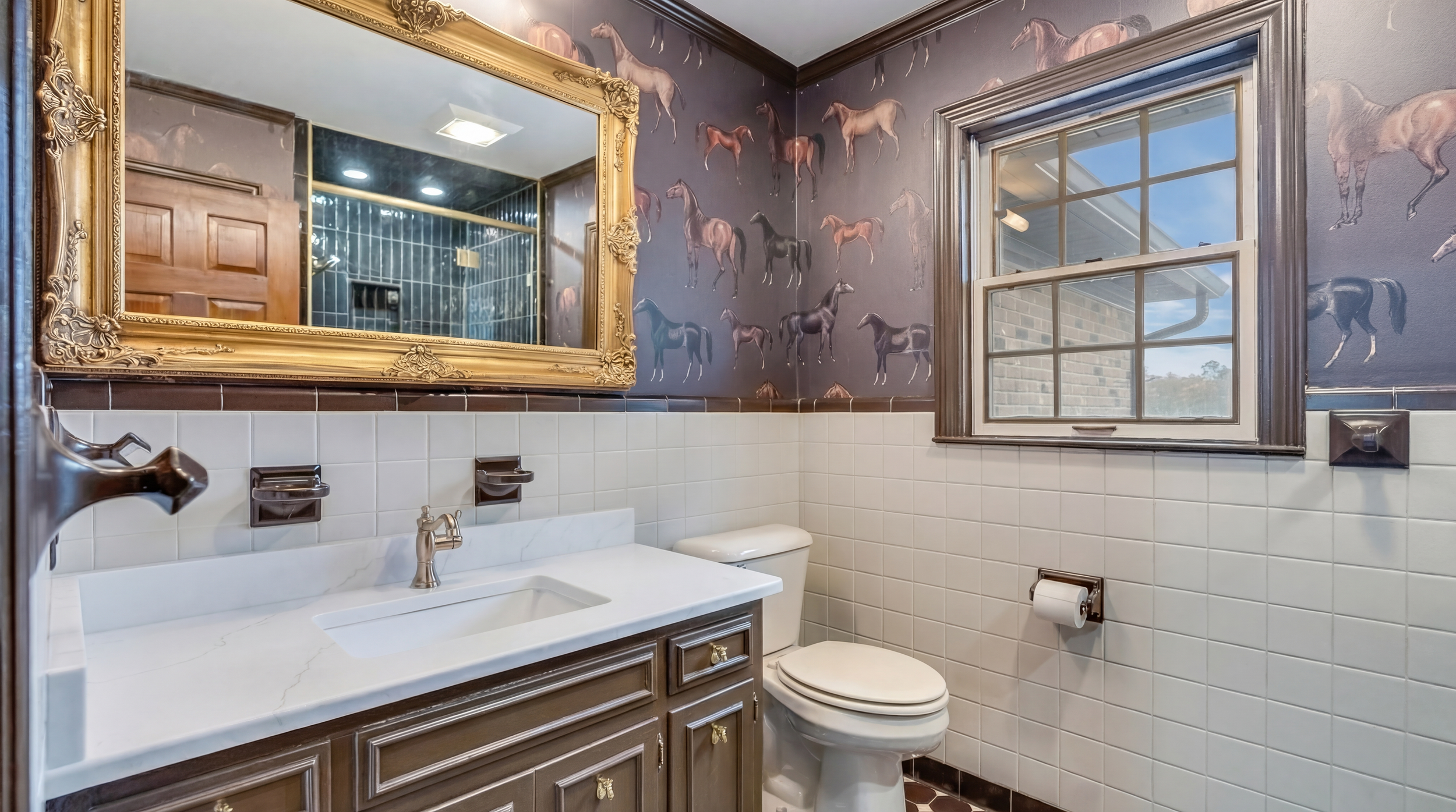 Bathroom with white tile halfway up the wall, wallpaper with horse pattern above, a wooden vanity with a marble countertop, large gold mirror, toilet, and window showing outdoors.