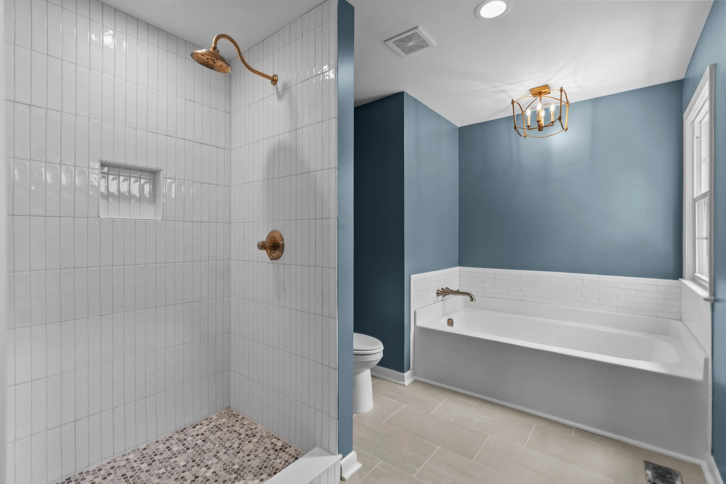A bathroom with a walk-in shower with white tile walls and a bronze showerhead, a separate bathtub with a bronze faucet, blue walls, a white window, and a ceiling light fixture.