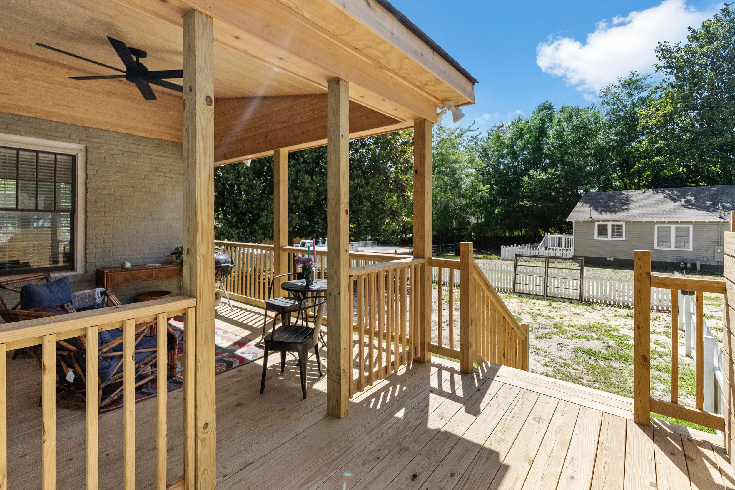 A wooden porch or deck attached to a house, with outdoor furniture including a small table, chairs, a sofa with cushions, and a ceiling fan. The porch has a staircase leading to a backyard with a fenced yard and trees.