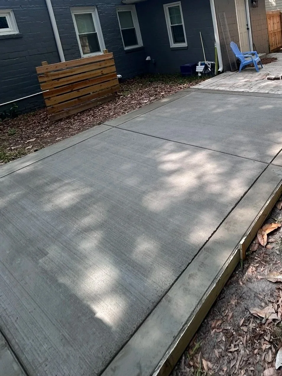 Freshly poured and smooth concrete patio or driveway in a backyard, bordered by a garden area with fallen leaves, next to a house with gray walls, two windows, outdoor chairs, and gardening tools.