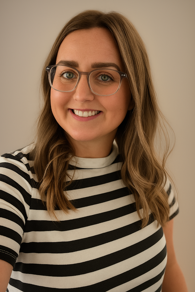 A young woman with light brown, wavy hair and glasses, smiling, wearing a black and white striped shirt, standing against a neutral background.