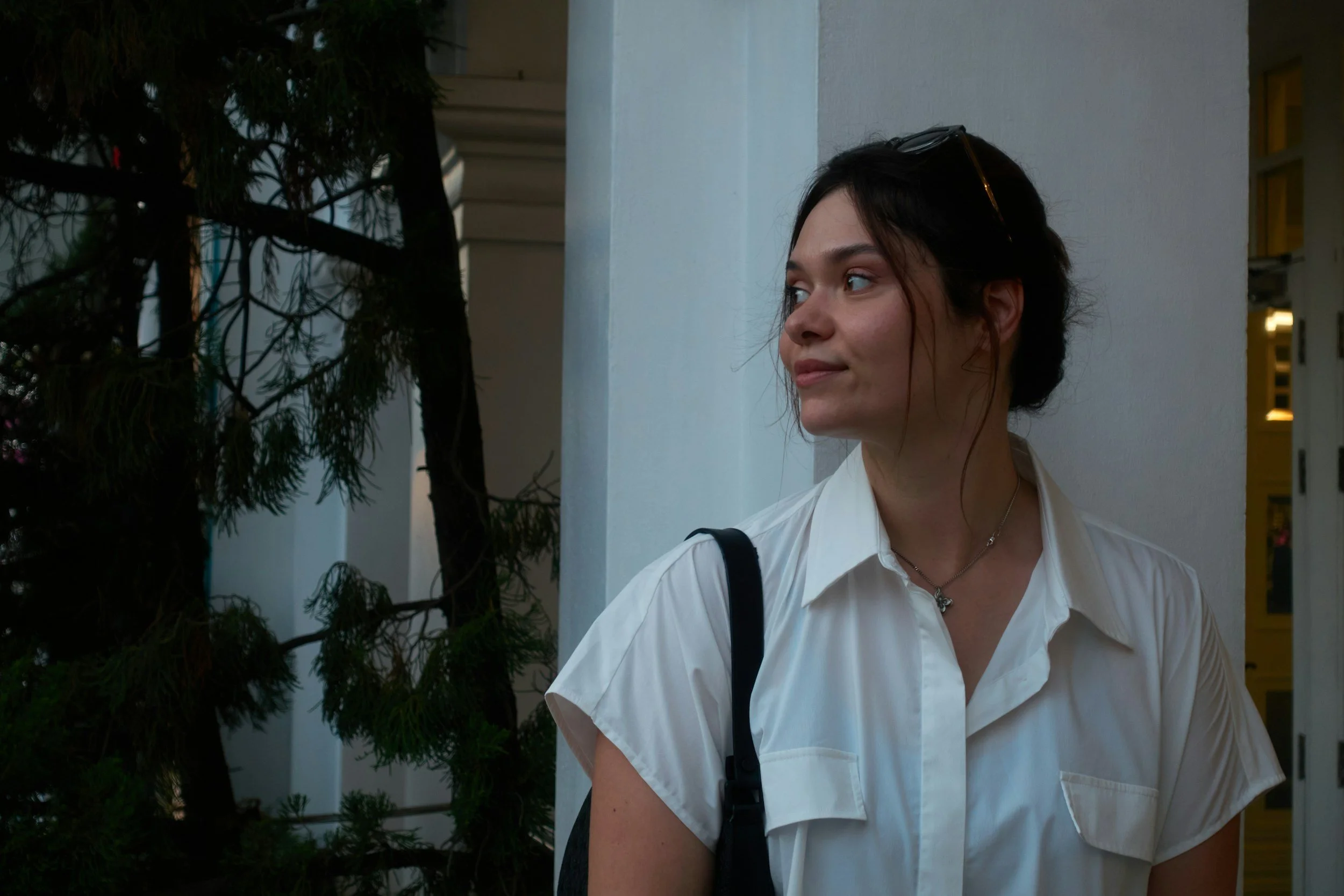 A woman with dark hair, wearing a white shirt and a necklace, standing outdoors near a white wall and some greenery, looking to her right.
