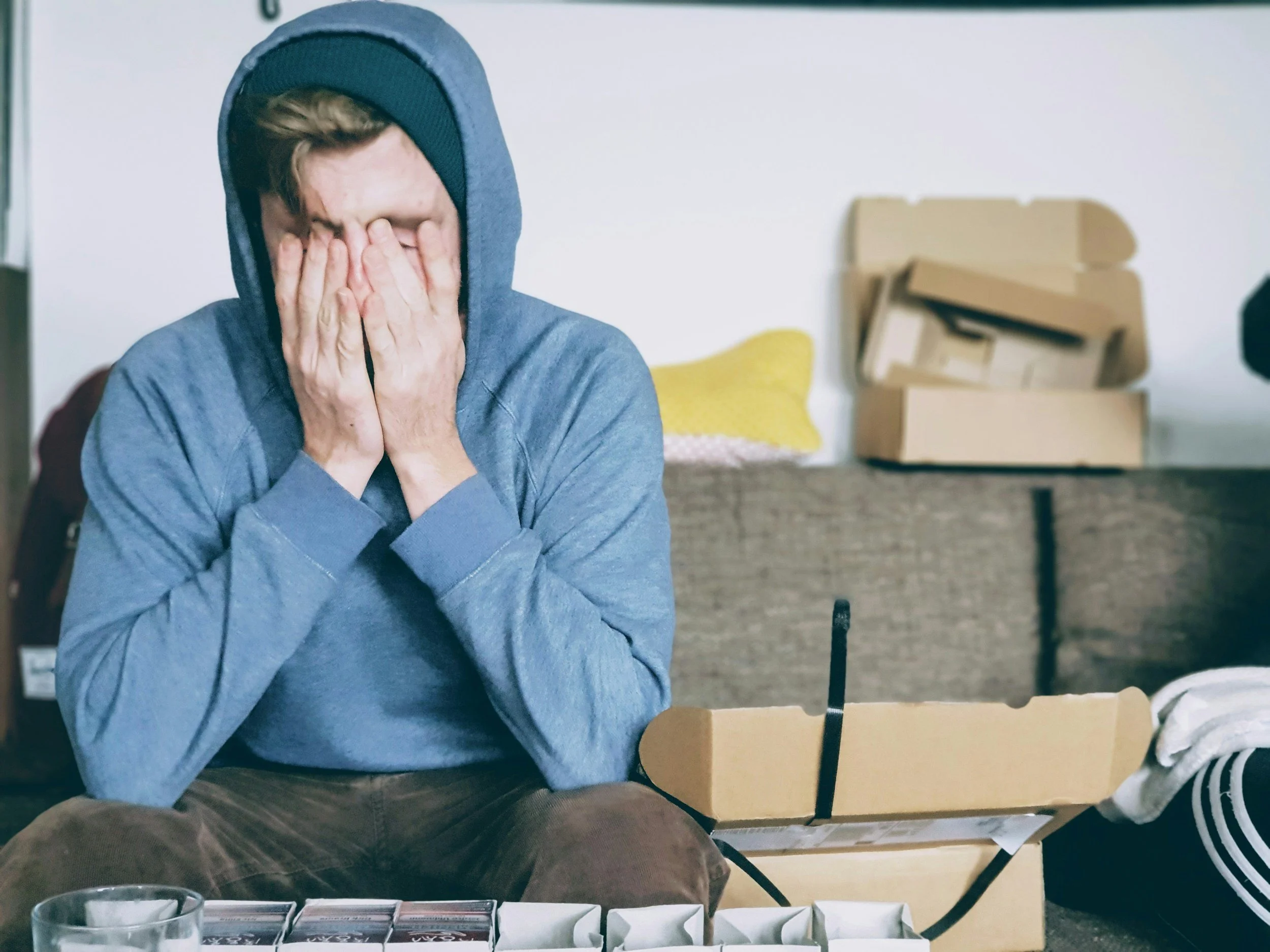 A man sitting on a couch wearing a hoodie and beanie with his hands covering his face, surrounded by empty boxes and closet items, indicating stress or sadness.