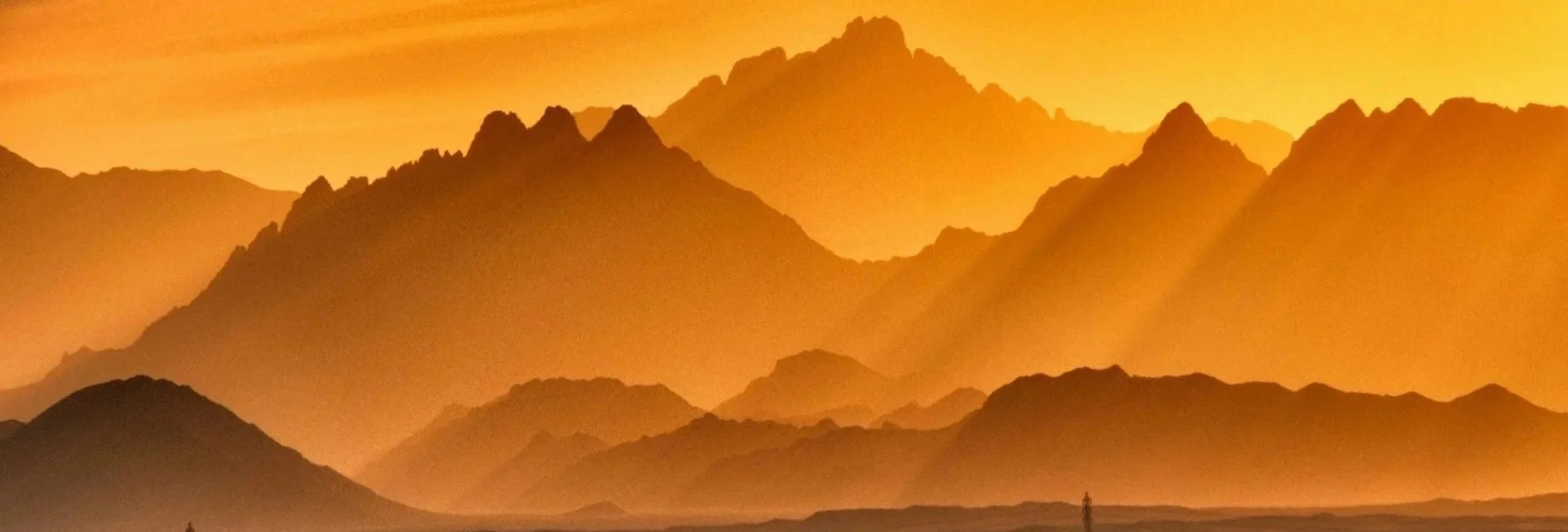 A landscape of layered mountains at sunset with an orange sky, and a person standing near the bottom right.