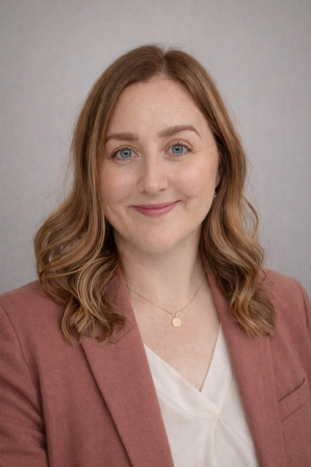A woman with red hair smiling, wearing a pink blazer and a white top with a gold necklace, against a gray background.