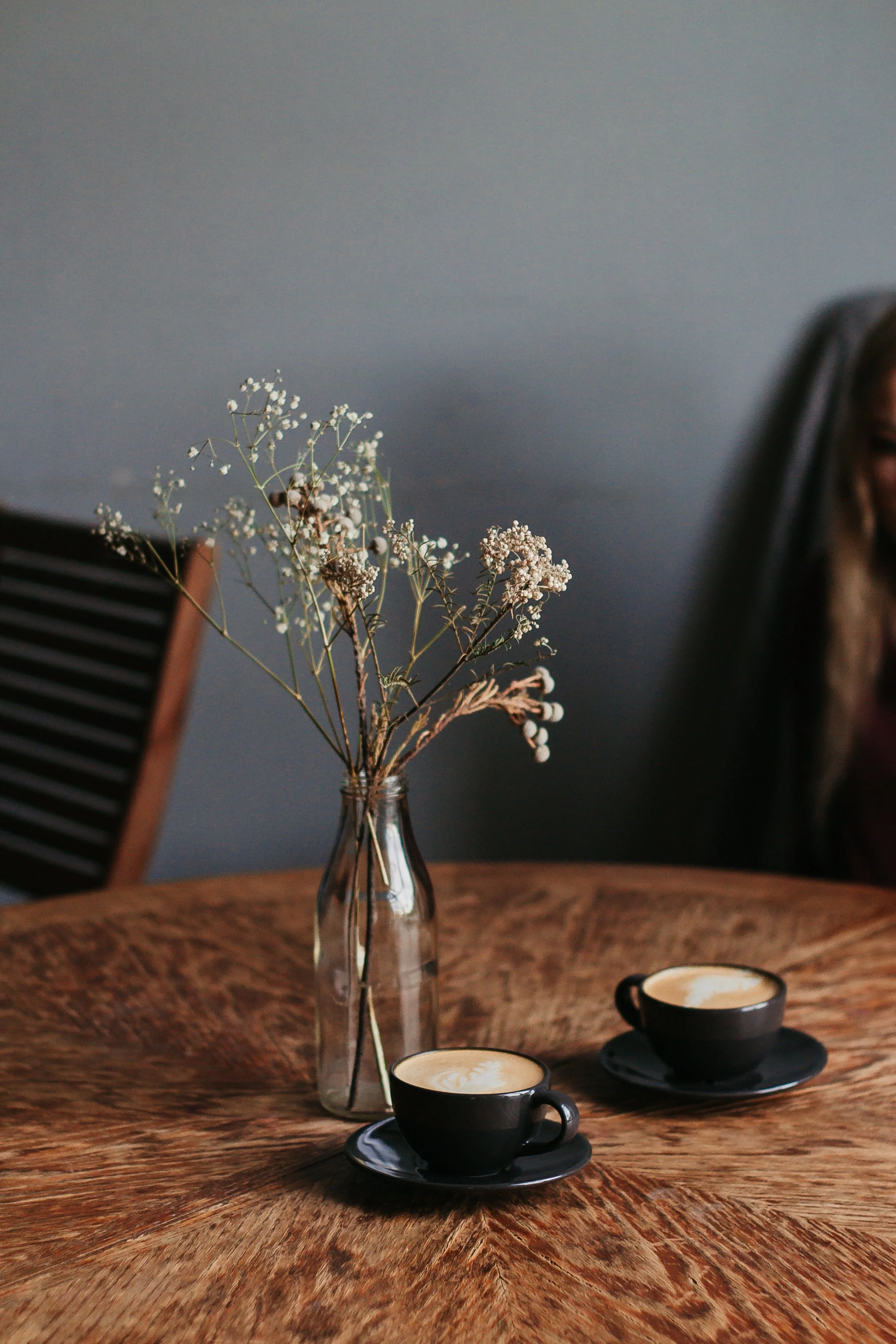 A wooden table with two black cups of coffee on black saucers, and a clear glass vase with dried white flowers, set against a gray wall.