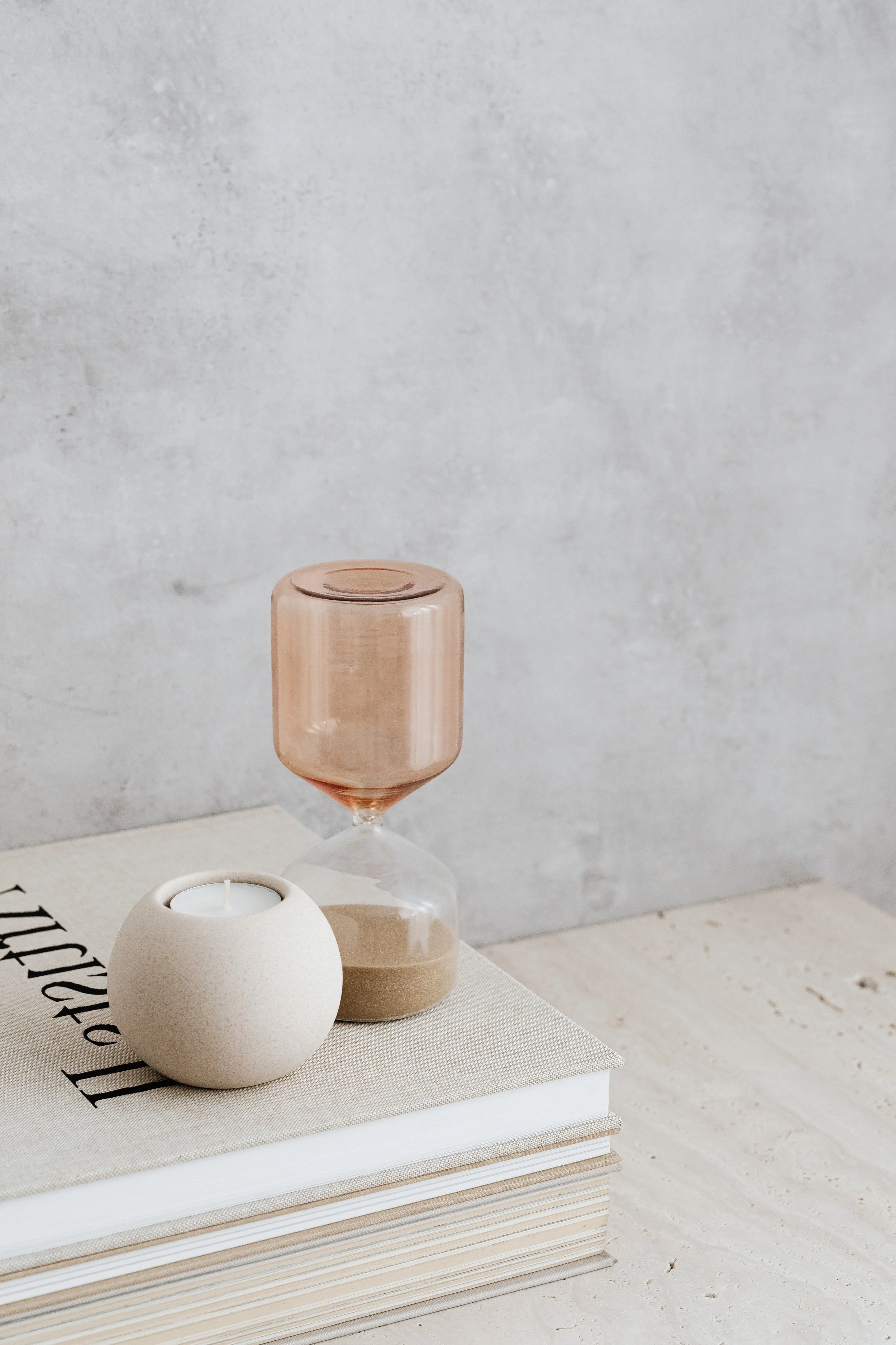 A beige candle holder with a candle inside, a pink-tinted glass hourglass, and a large book with a light-colored cover on a wooden surface against a gray wall.