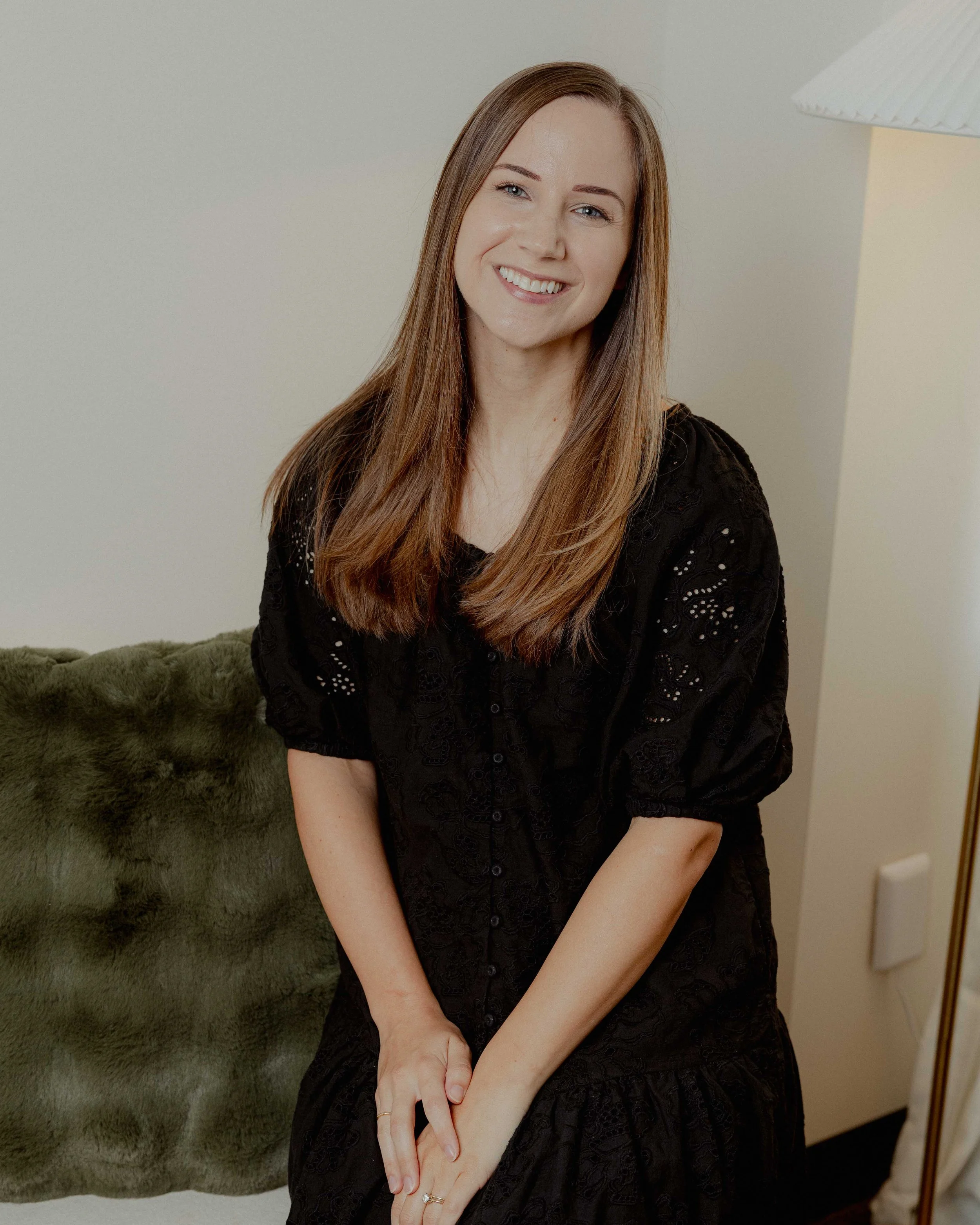 Portrait of a smiling woman with long auburn hair, wearing a black lace dress, sitting with her hands clasped on her lap in front of a plain wall and a lamp.