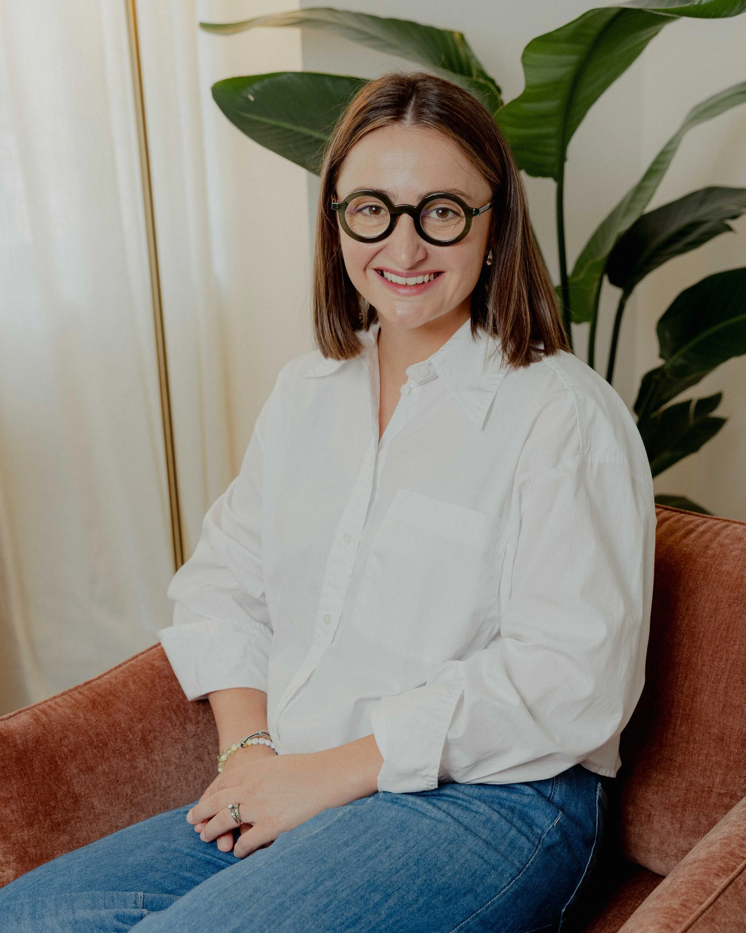 A woman with shoulder-length brown hair, wearing large round black glasses, a white button-up shirt, and blue jeans, is sitting on a reddish-brown couch, smiling at the camera with her arms crossed, in a room with large green plant leaves behind her.