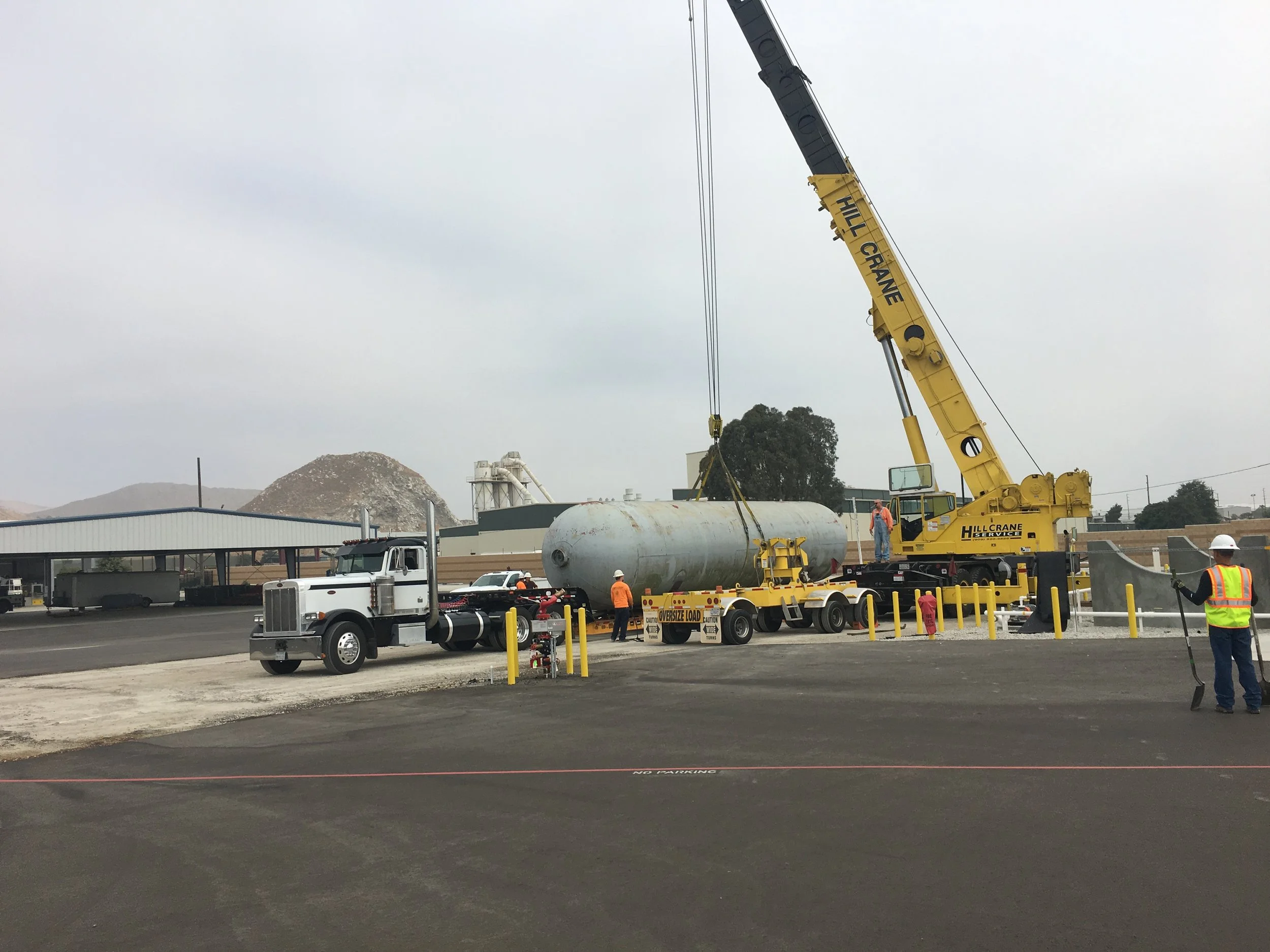 Construction site with a yellow crane lifting a large cylindrical metal tank. Several workers in safety vests and helmets are present, and trucks are parked nearby. The sky is overcast.