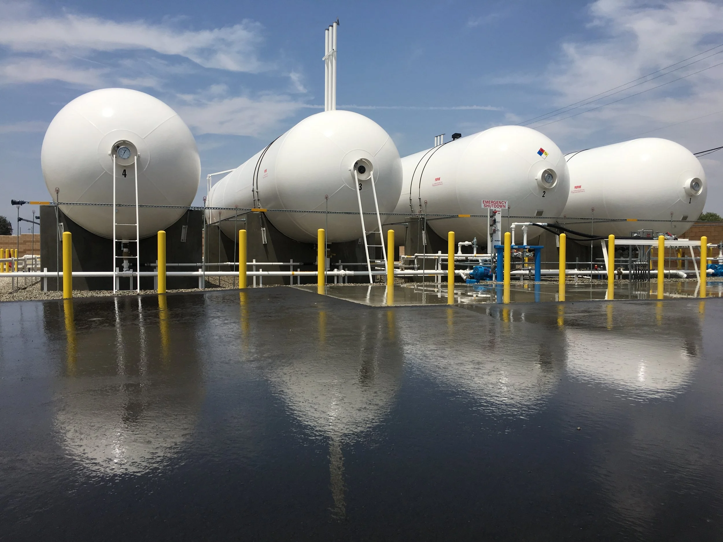 Multiple 30,000 gallon liquid propane storage tanks with yellow bollards and painted manifold piping on a newly surfaced yard.