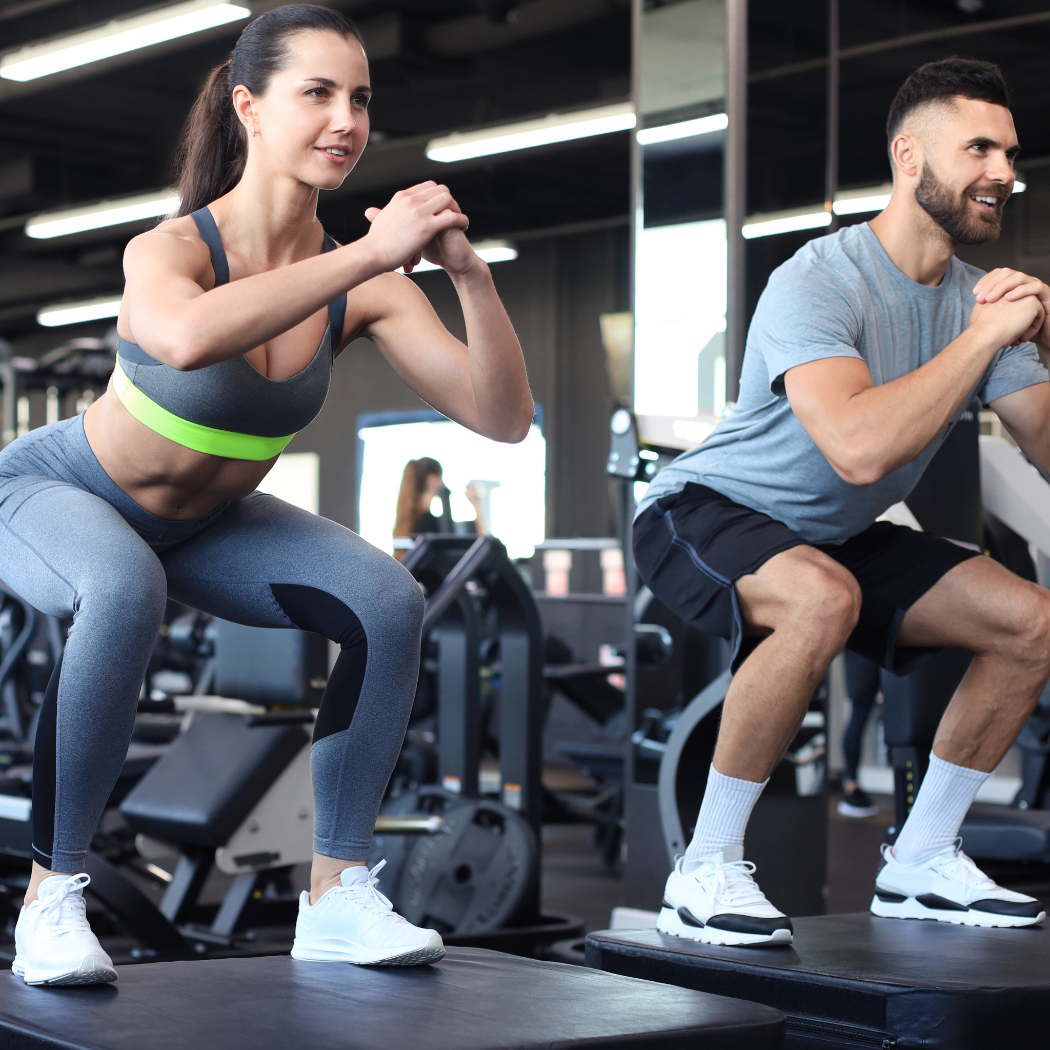 Two people performing squats on exercise platforms at a gym, with fitness equipment in the background.