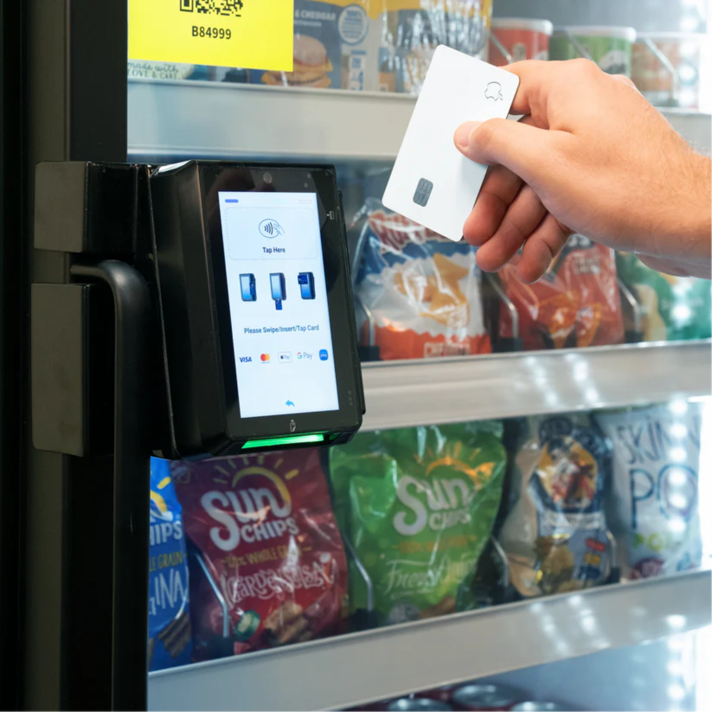 A person is holding a white Apple Pay card near a contactless payment terminal at a grocery store, with shelves of snack bags in the background.
