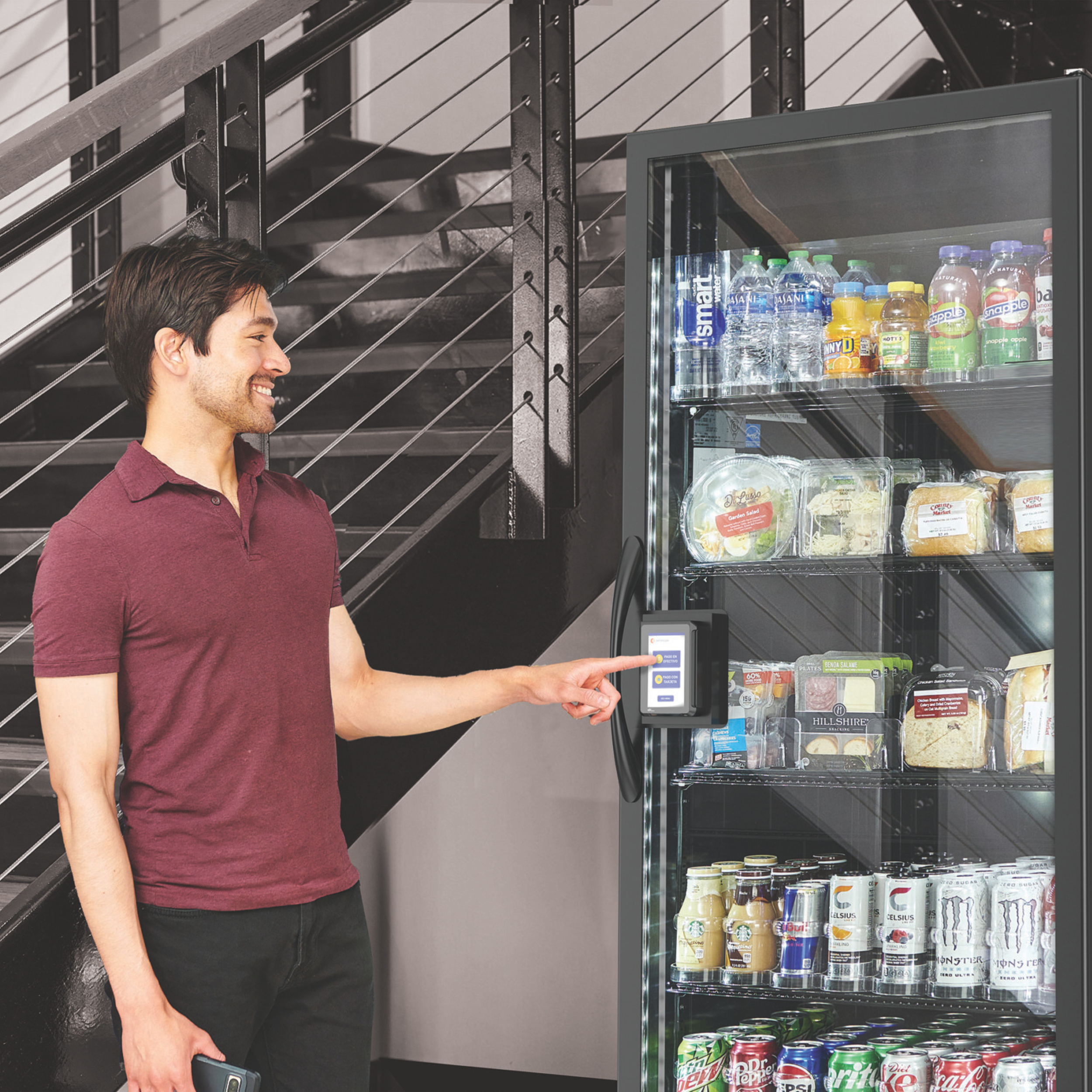 A man in a maroon polo shirt and black pants standing next to a vending machine with a touchscreen, selecting a snack or beverage, near a staircase with black metal railings.