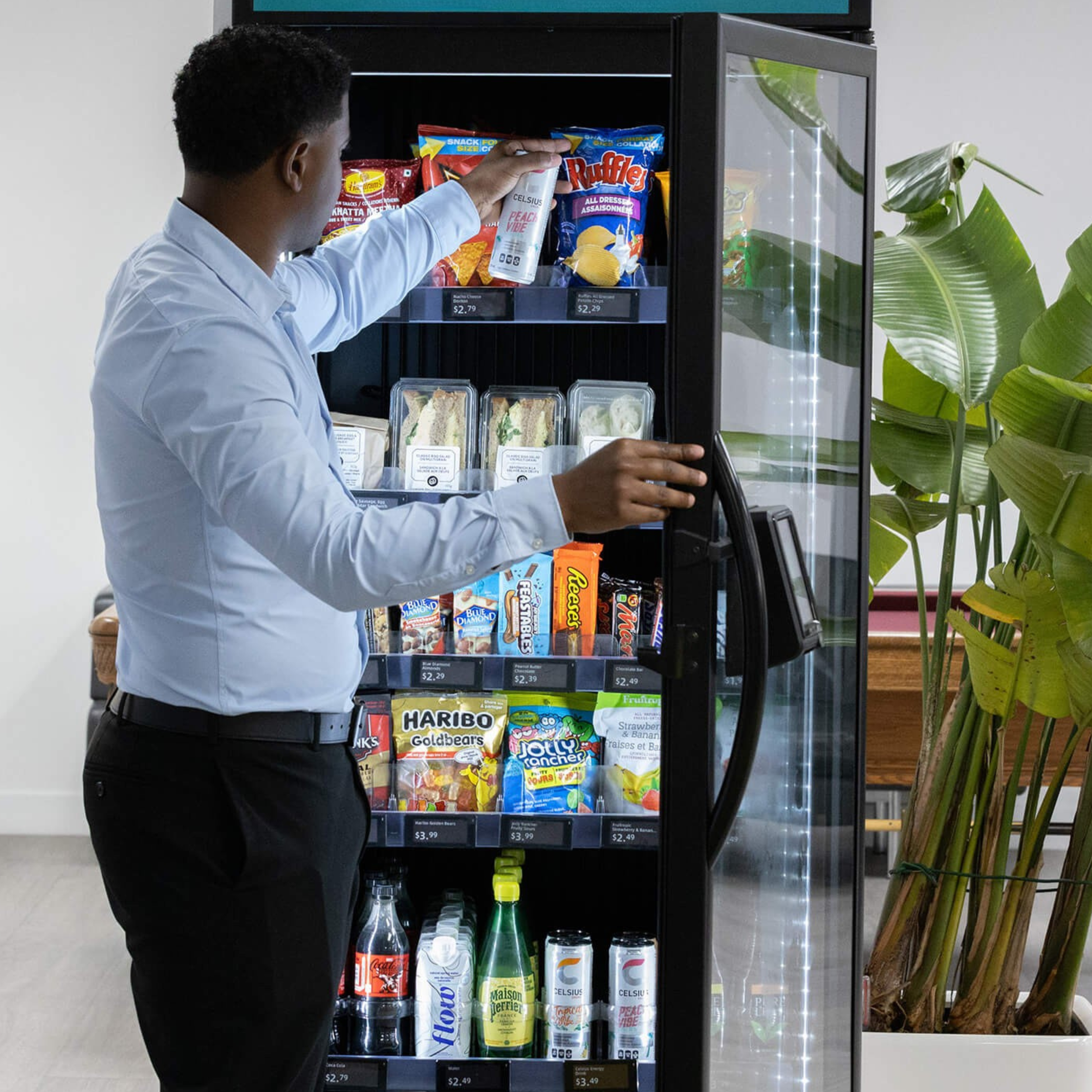 A man in a white shirt and black pants is choosing snacks from a vending machine, which contains chips, sandwiches, candy, and drinks, with green plants nearby.