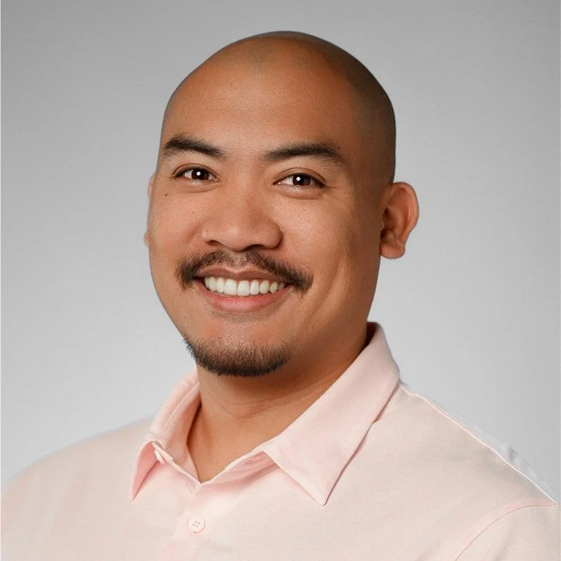 A smiling man with a shaven head, mustache, and goatee, wearing a light pink collared shirt, against a plain gray background.